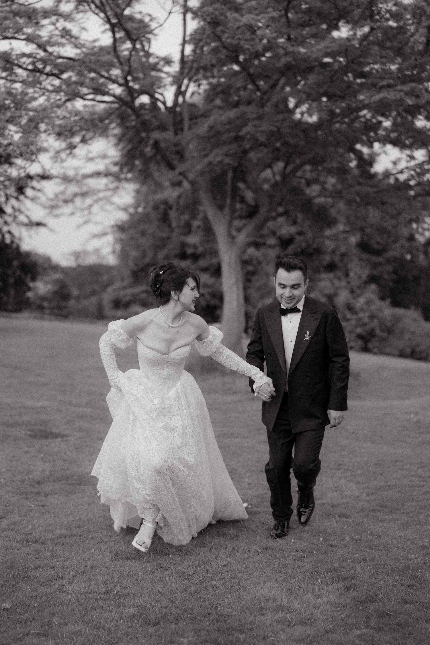 A black and white photo of a couple walking together on grass, with her dress flowing in motion.