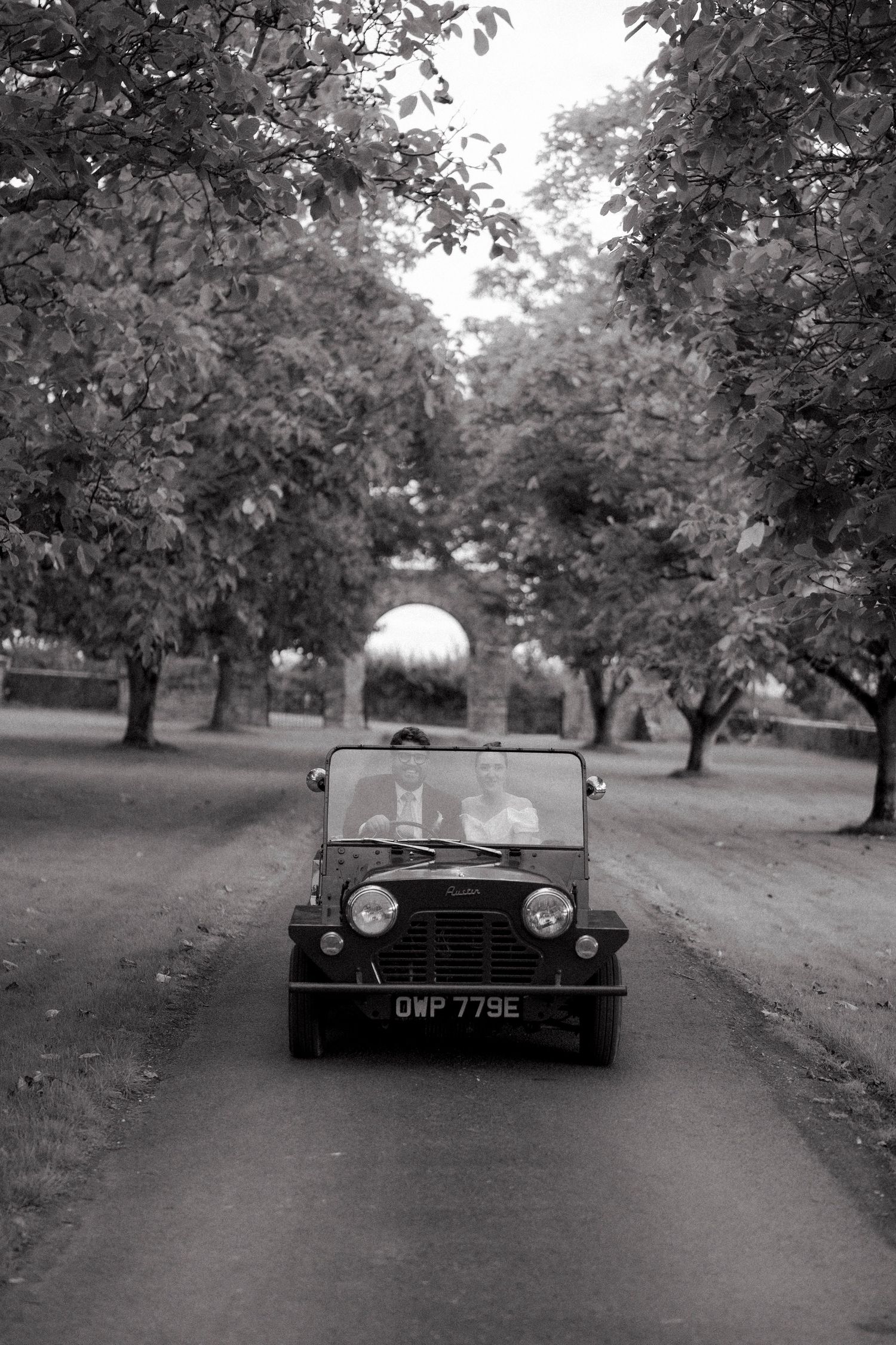Black and white photo of bride and groom driving a vintage Mini Moke down a tree-lined driveway.