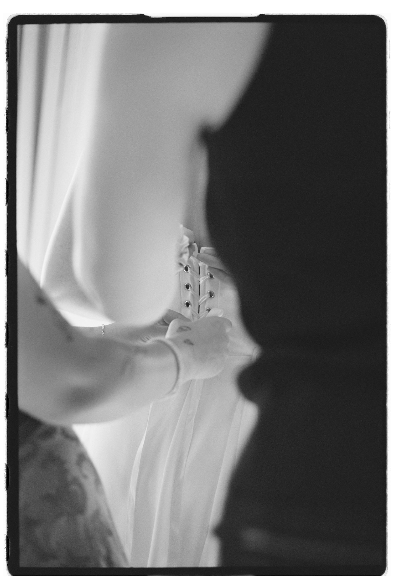 Black and white photo of a bride being helped into her wedding dress, showing delicate button details on the back.