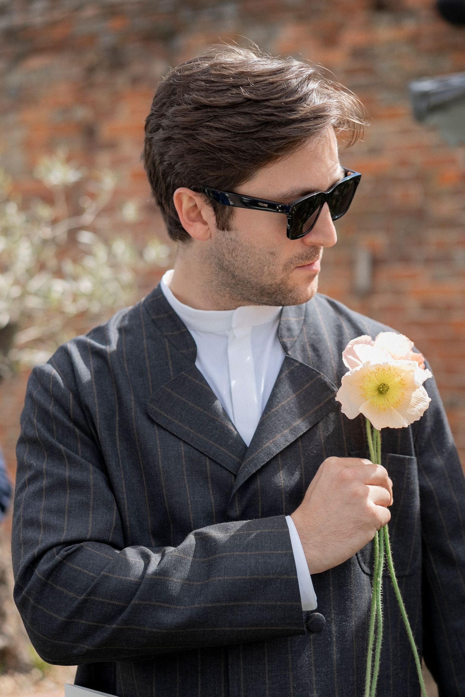 Man in sunglasses and a pinstripe jacket holds a pale pink poppy flower in front of a brick wall.