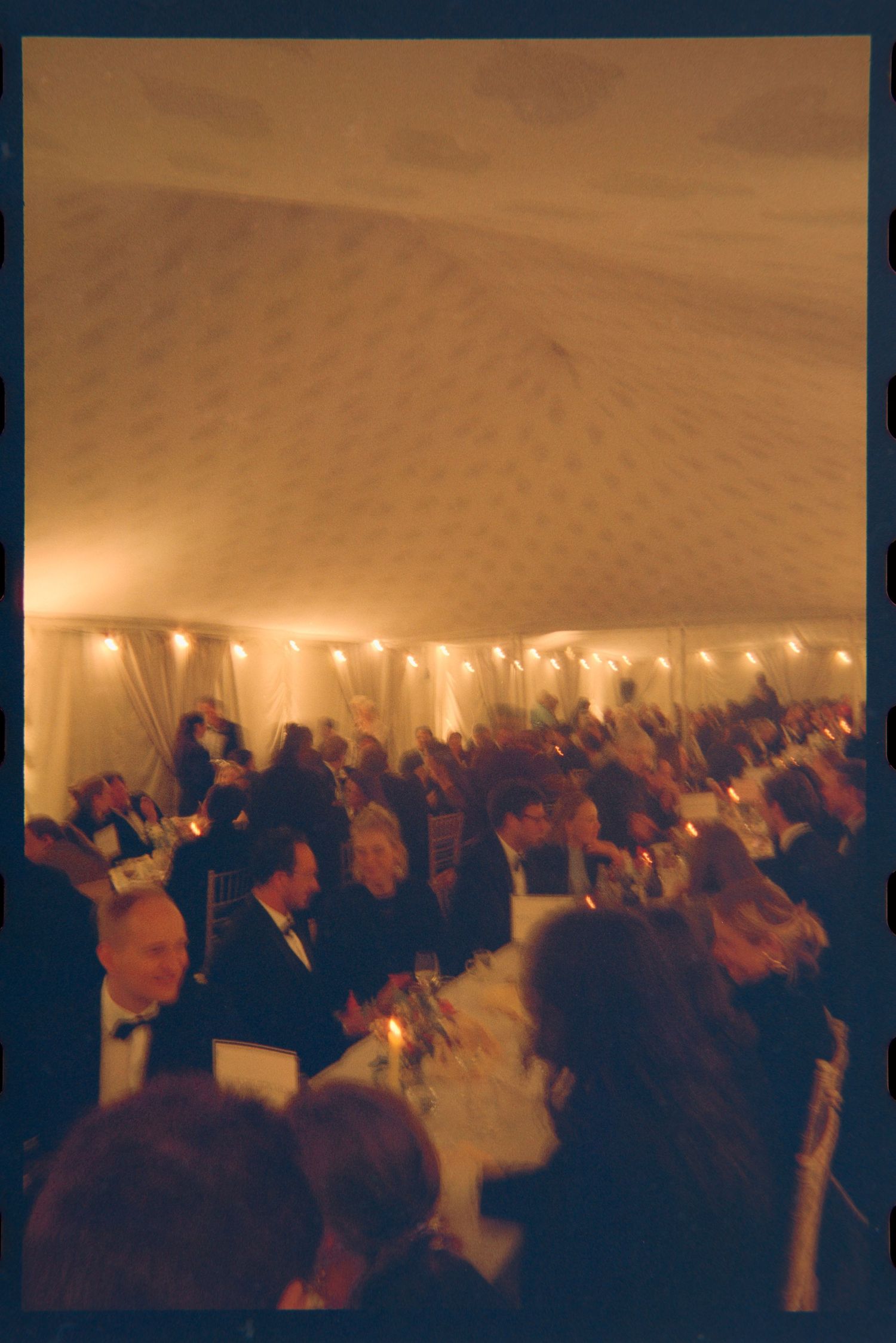 Wedding guests in formal attire dine at long tables inside a warmly lit marquee tent with a draped ceiling and glowing string lights.