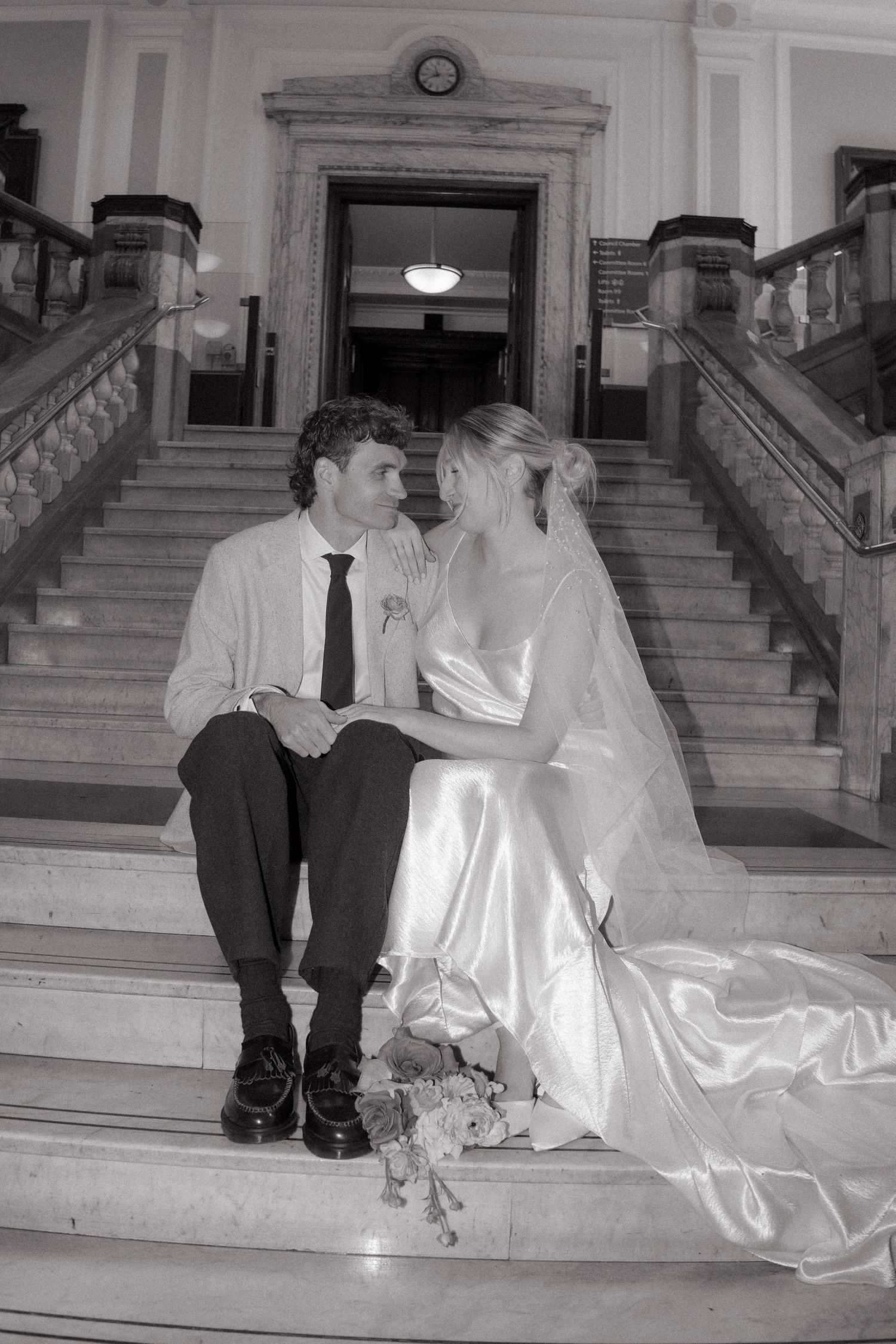 Black and white photo of a bride and groom sitting on marble stairs, sharing a romantic moment in wedding attire.