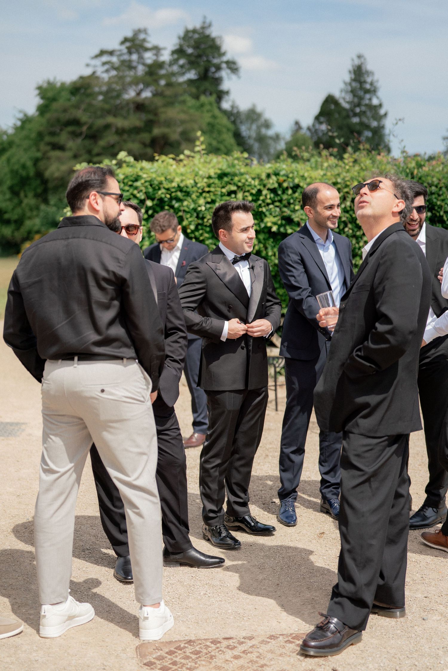 Wedding guests in dark suits and casual attire mingling outdoors during a reception.