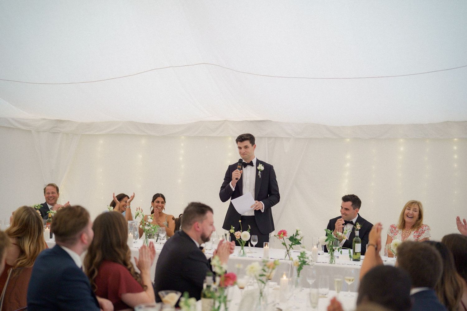 Wedding reception guests gathered inside an elegant white tent with floral centerpieces on tables.