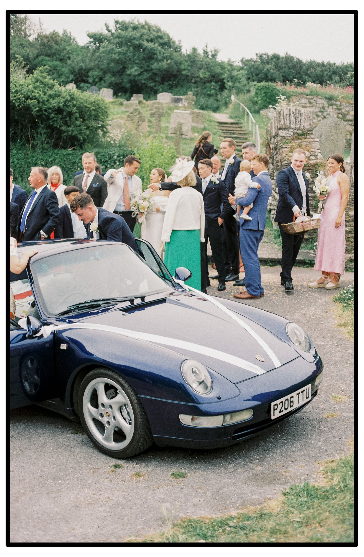 A sleek navy blue Porsche sports car parked at an outdoor event with guests gathered around.