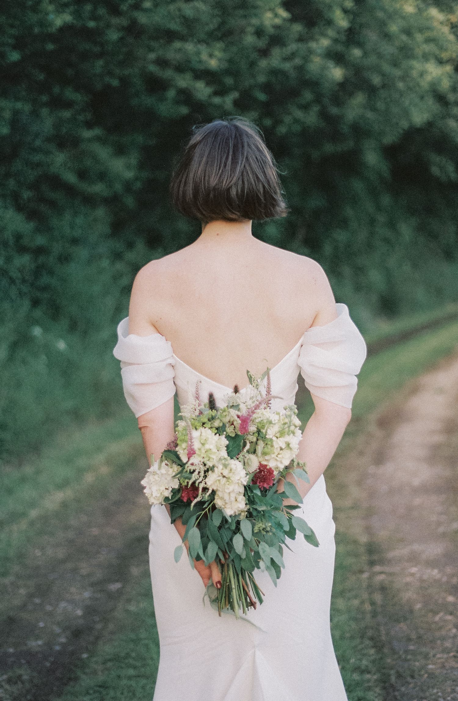 A bride in a white off-shoulder wedding dress holds a romantic bouquet of pink and cream flowers on a forest path.