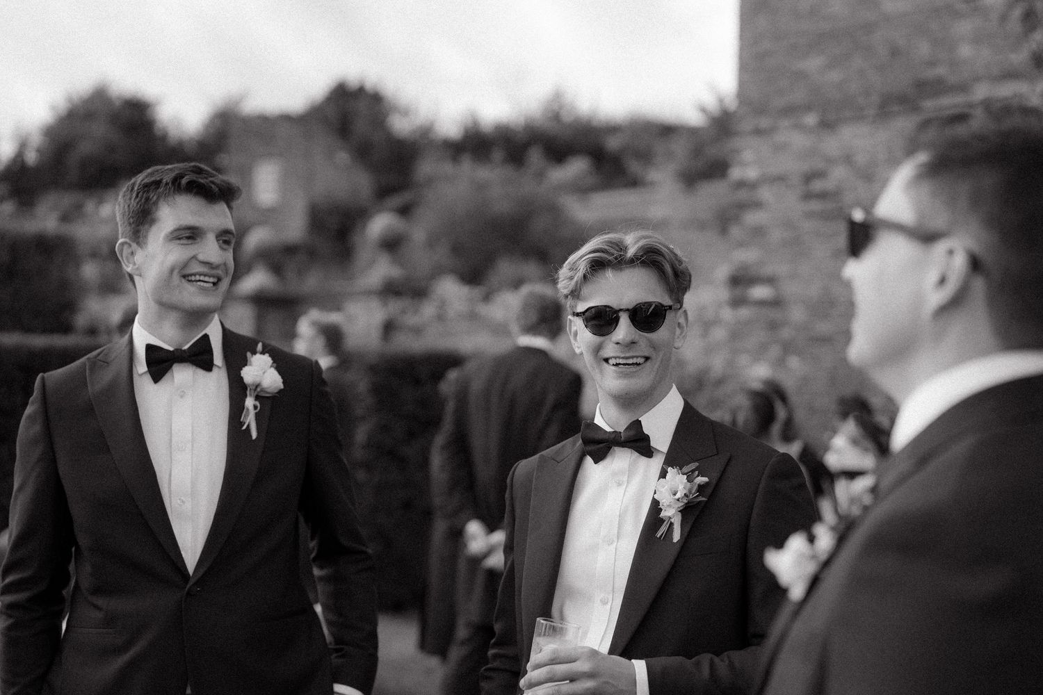 Wedding guests in formal attire and sunglasses gathered outdoors in black and white photography.