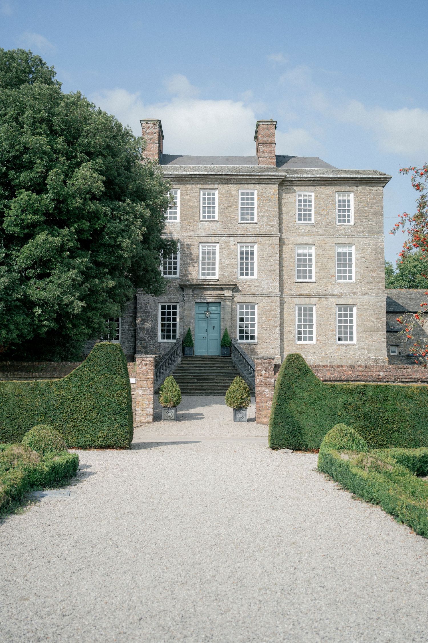 Georgian country house with gravel driveway, manicured hedges, and teal front door under a blue sky.
