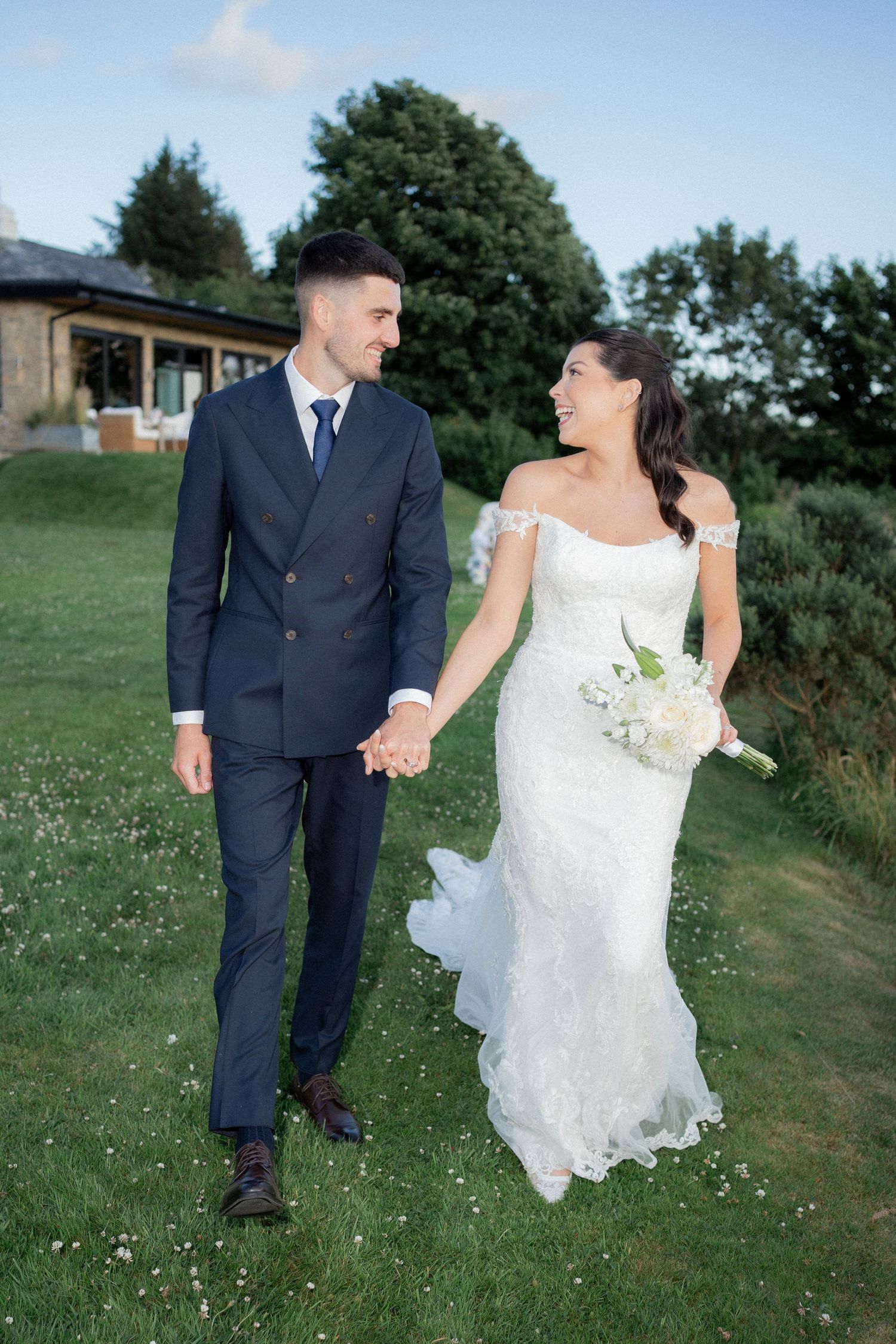 A newlywed couple walking hand in hand on grass with a venue in the background.