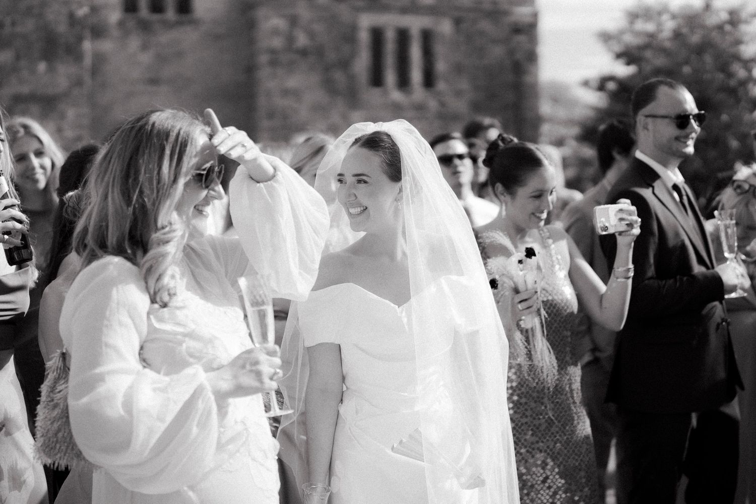 Smiling bride in white dress and veil chatting with guests at outdoor wedding reception.