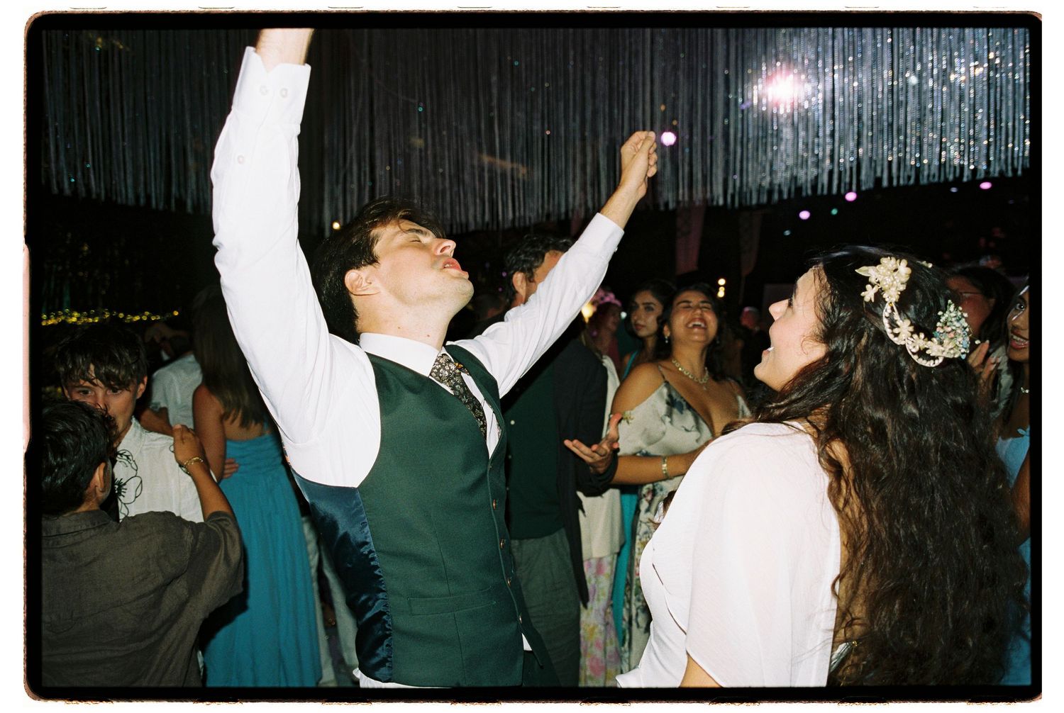 Groom raises arm joyfully on wedding dance floor while bride smiles beside him at elegant reception.