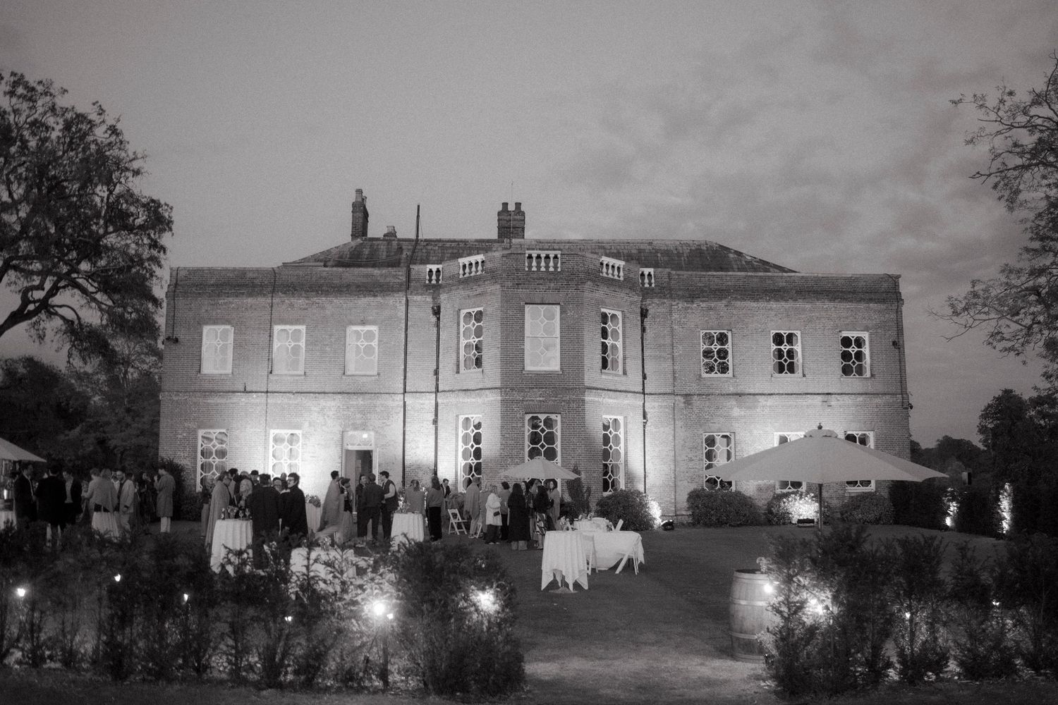 Black and white photo of guests gathering outside an illuminated Georgian manor house at dusk during an elegant outdoor wedding reception.