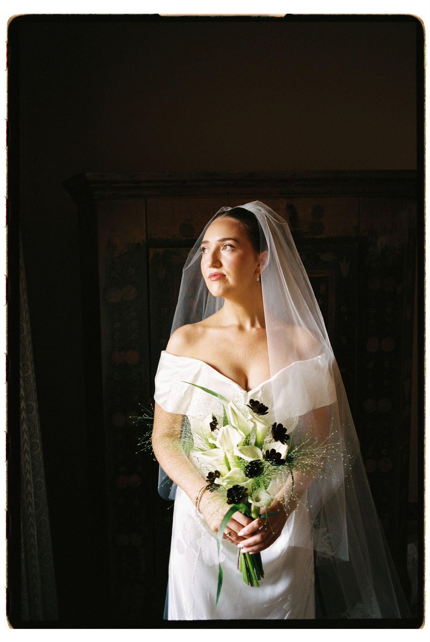 Bride in white off-shoulder gown and veil holds white and black floral bouquet, gazing softly in natural light.