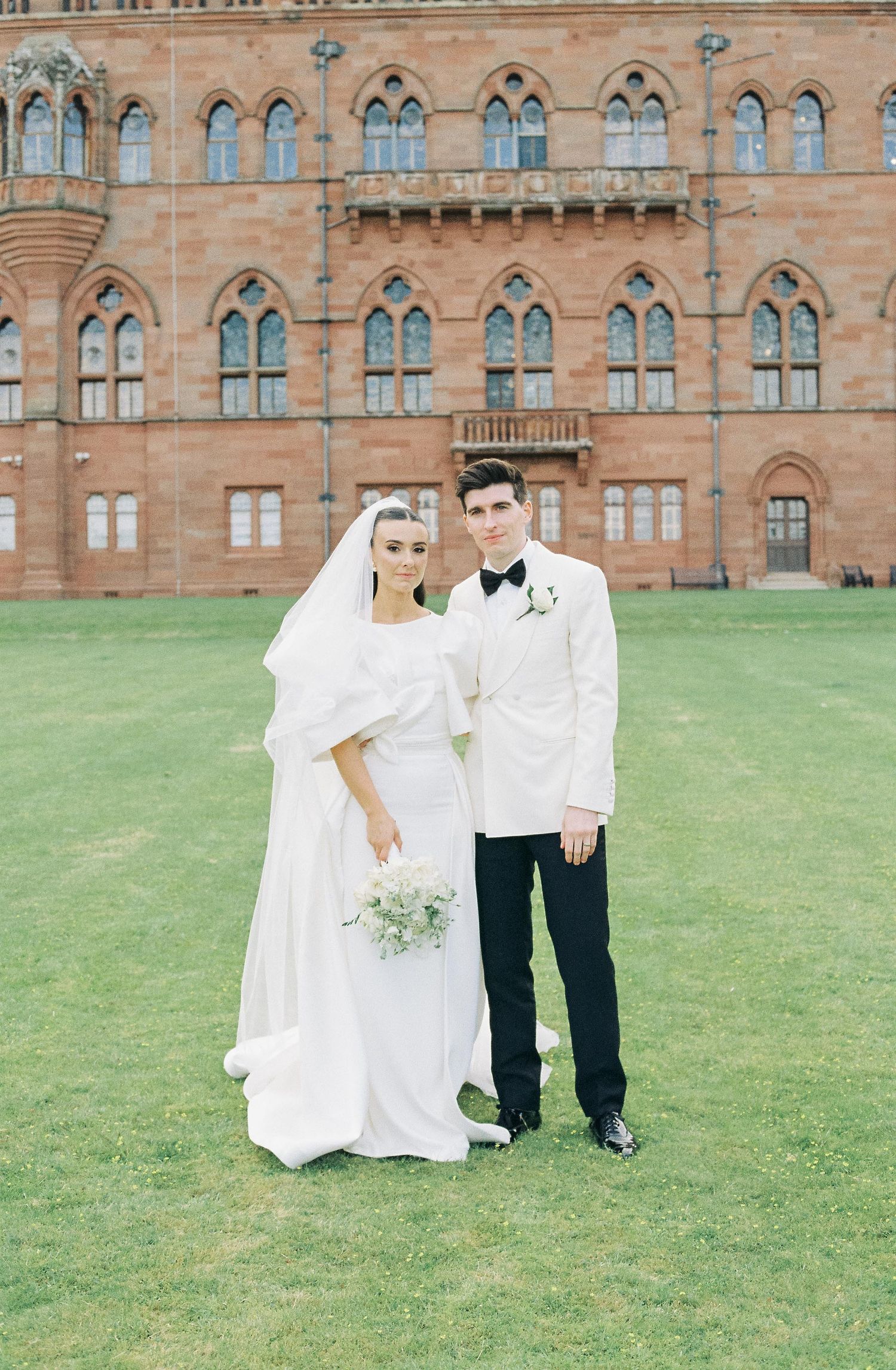 Wedding couple in white attire posing in front of historic Gothic revival building.