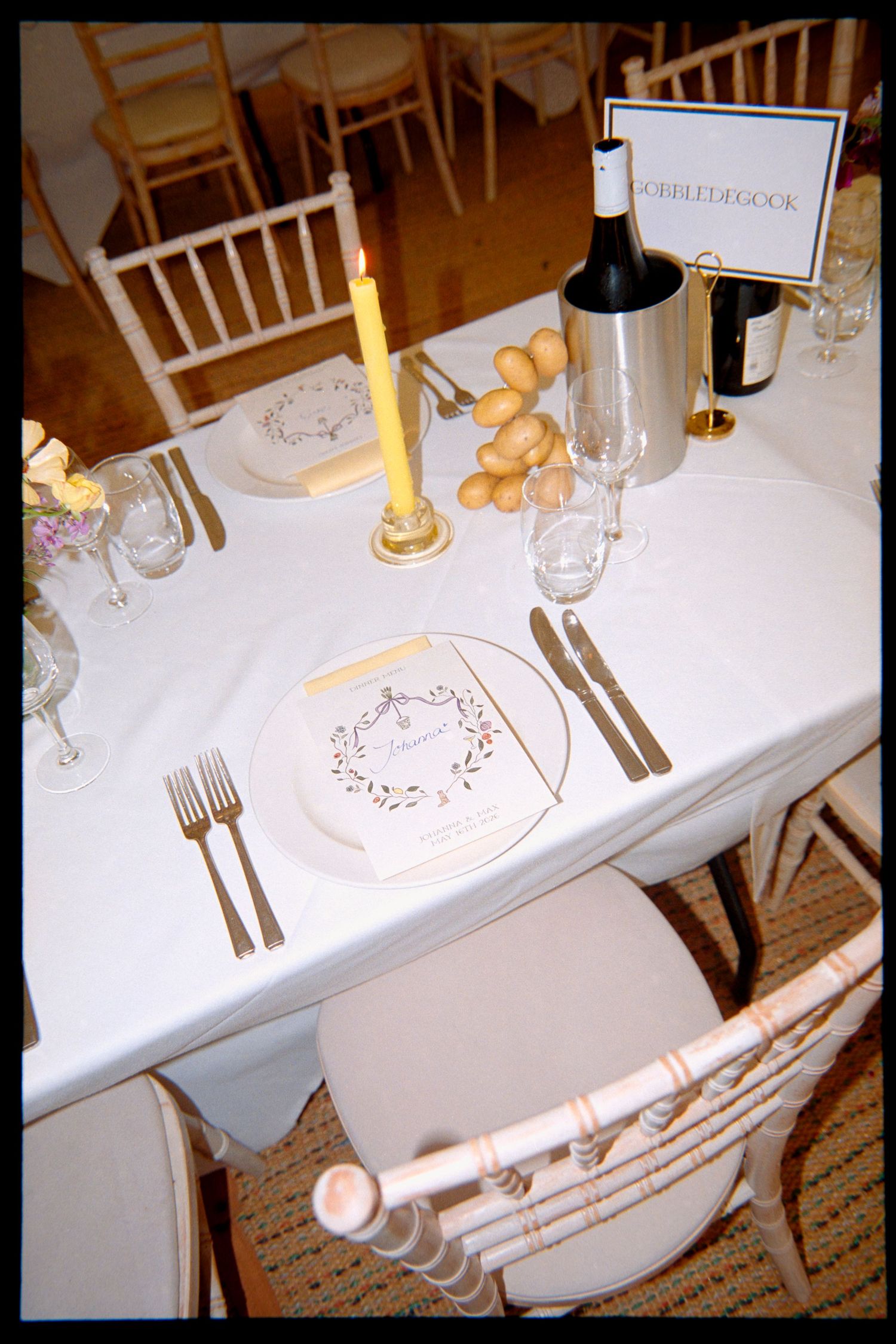 Elegant wedding table setting with printed menu, gold candle, wine bucket, glassware and white chairs.