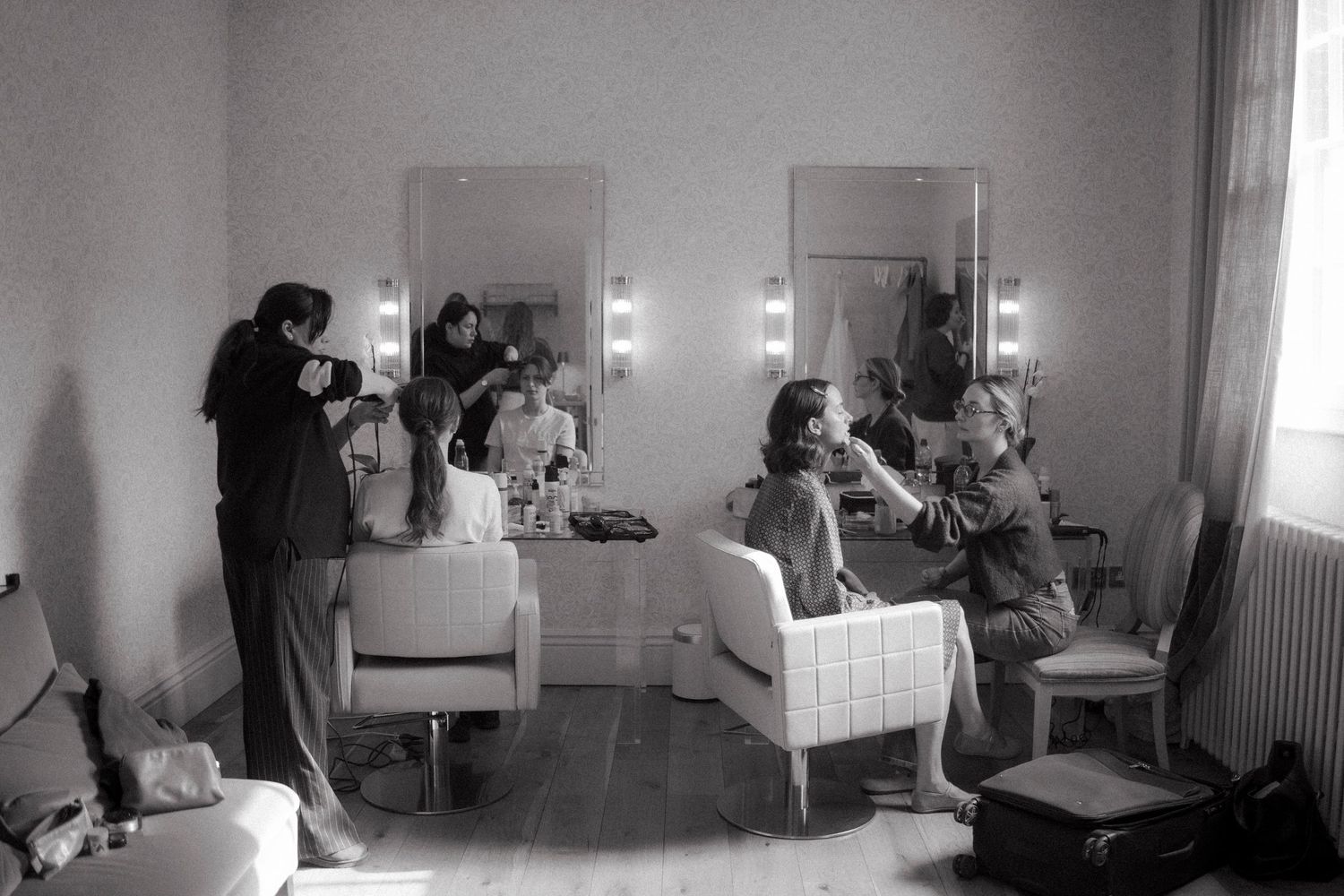 Black and white photo of women getting ready in a vintage dressing room with lit mirrors, while a photographer captures the moment.