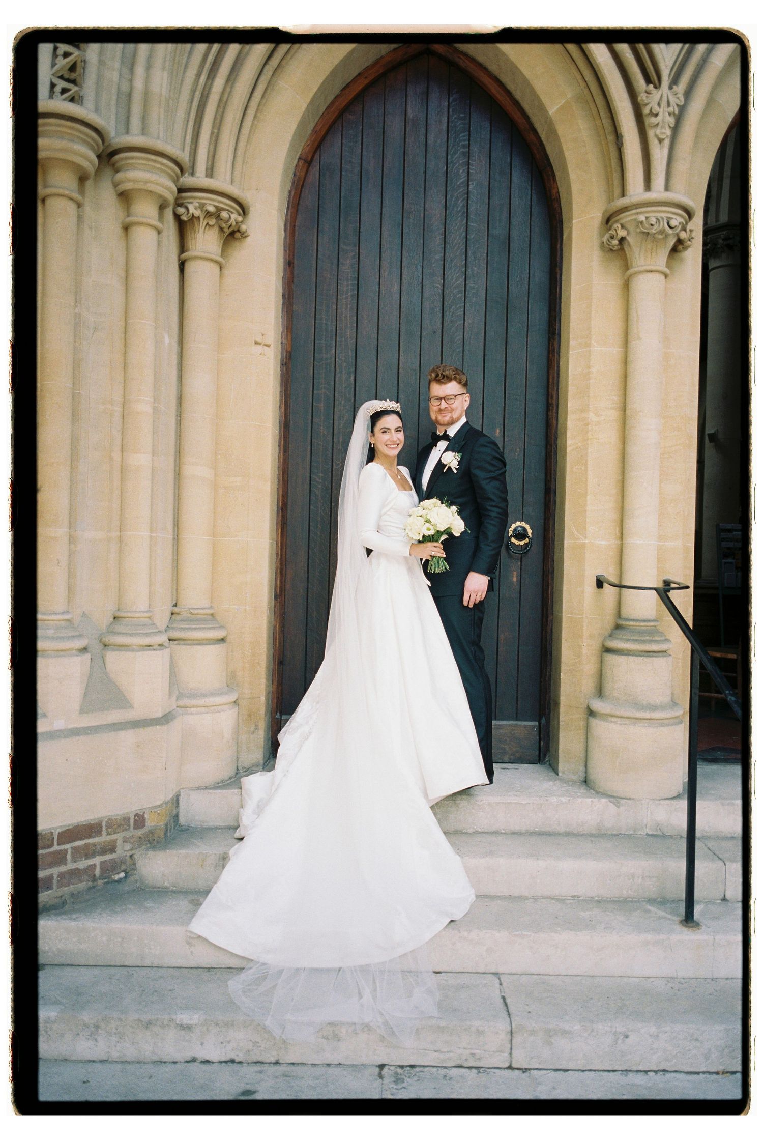 Bride and groom posing on stone steps before grand Gothic church wooden arched doorway with bouquet.