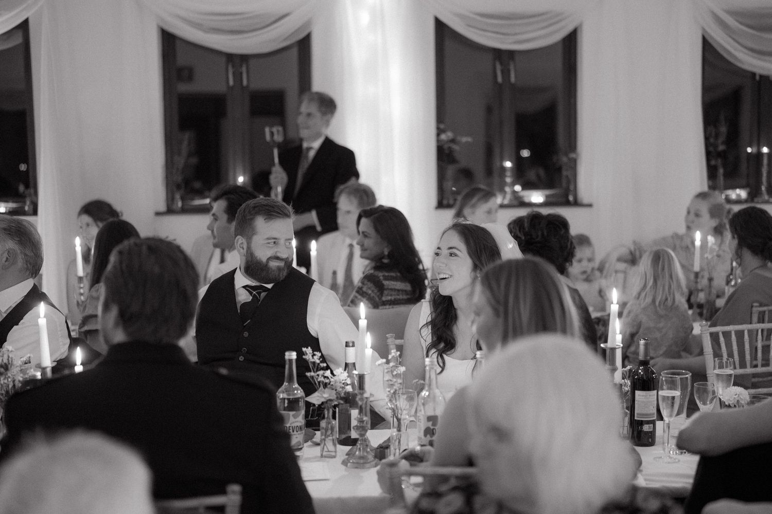 Black and white photo of guests enjoying a formal dinner reception at an elegant venue with white draped decor.