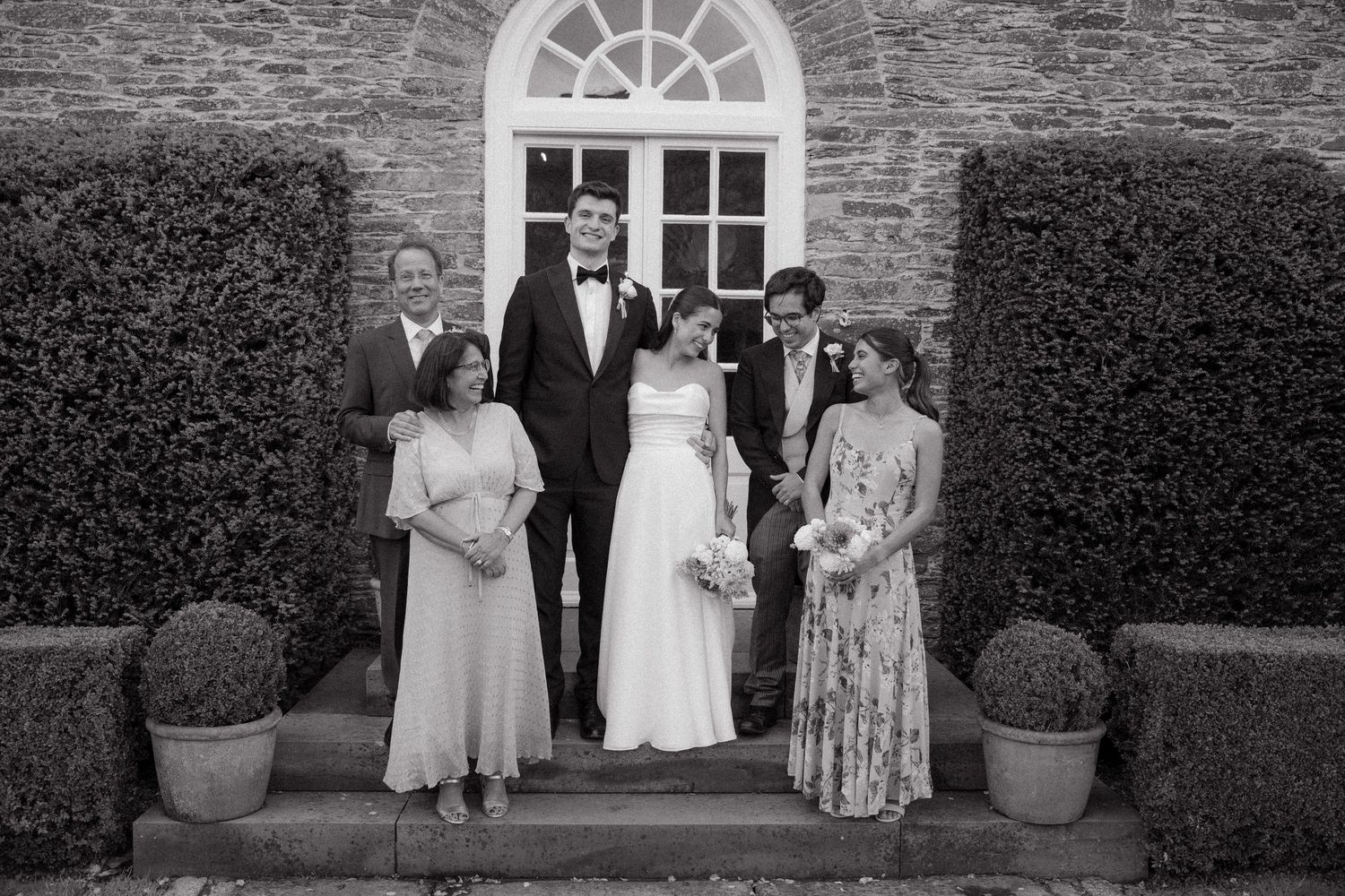 Stone building exterior with white-framed windows and manicured hedges during a wedding celebration.
