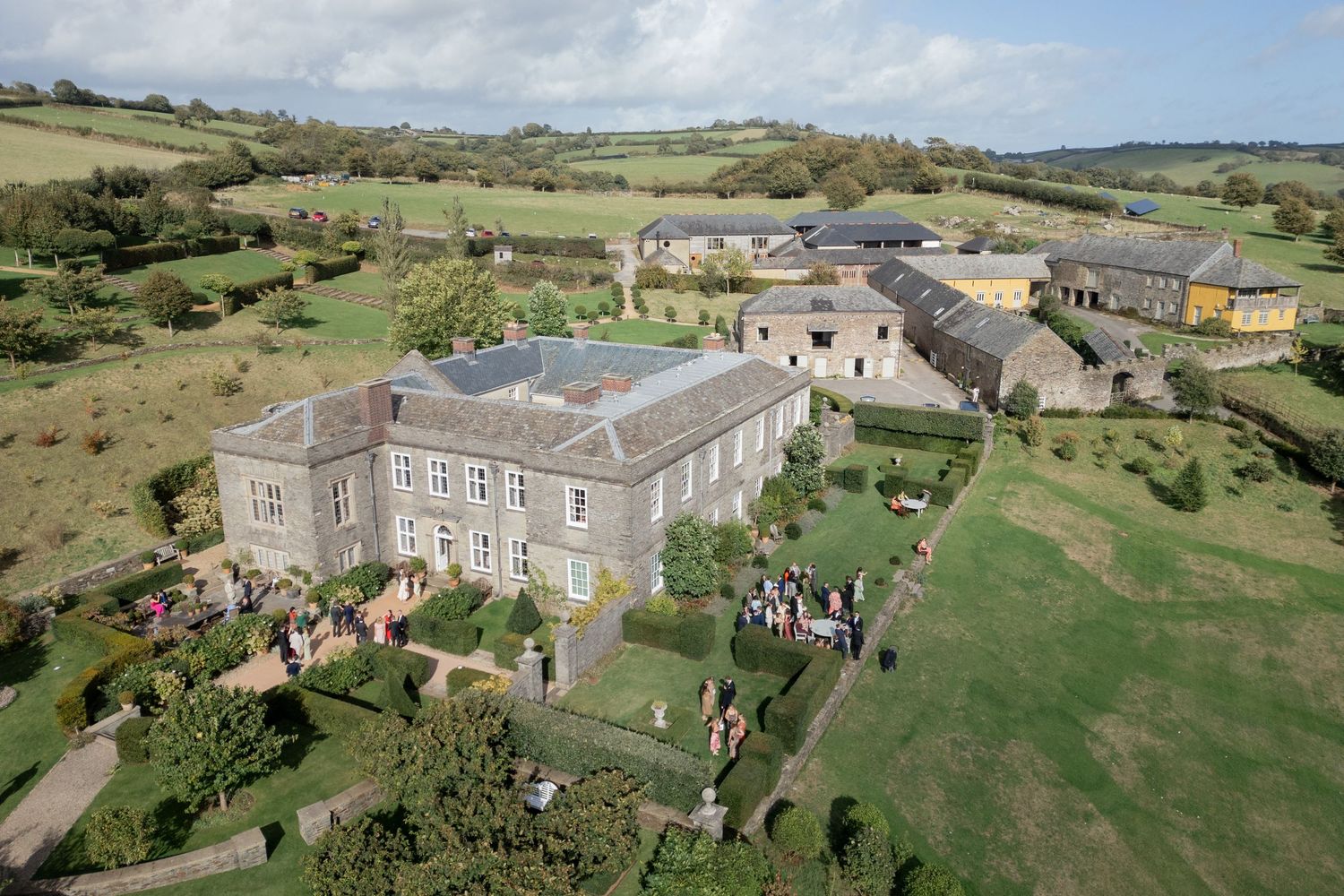 Aerial view of a grand country estate with manicured gardens, guests gathered outside, surrounded by green rolling fields.