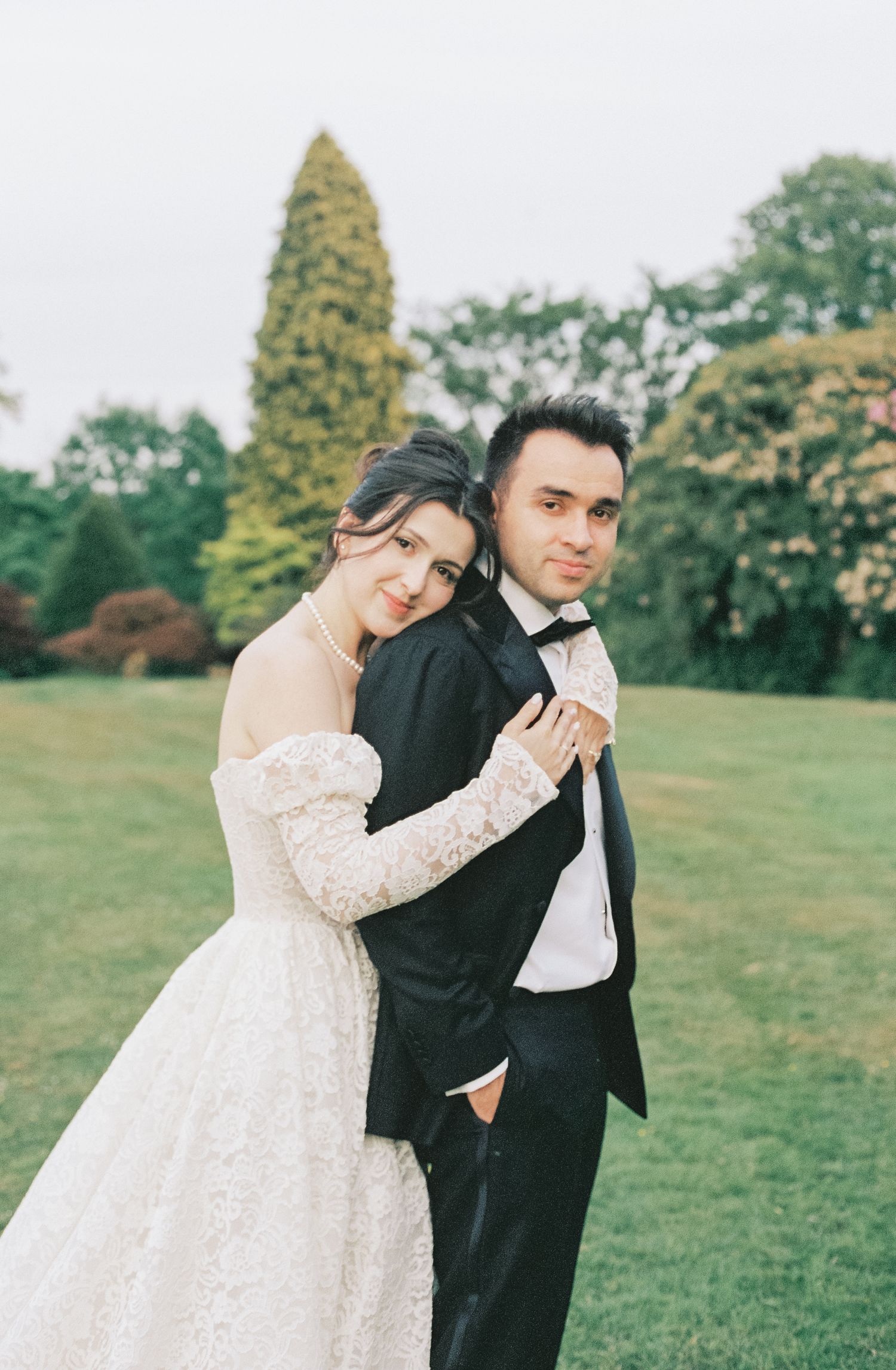 A romantic wedding couple embraces on a lush green lawn with trees in the background.