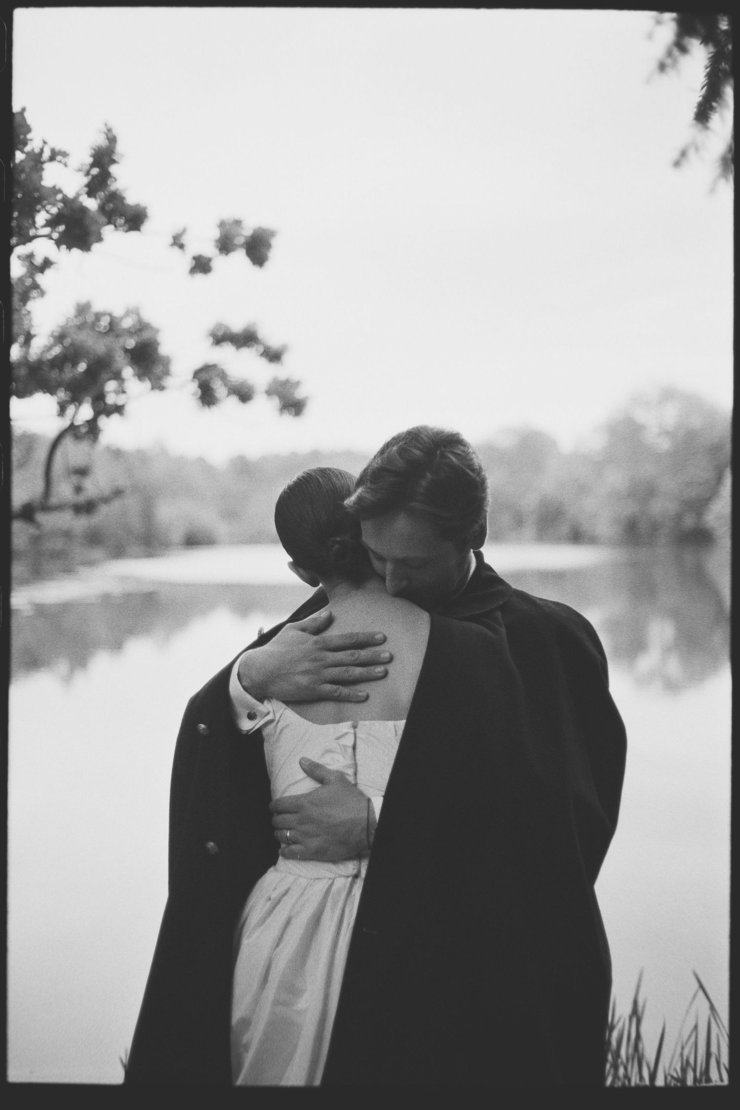 Black and white photo of a couple embracing by a calm lake, the woman in a white dress and the man in a dark coat, trees in the background.