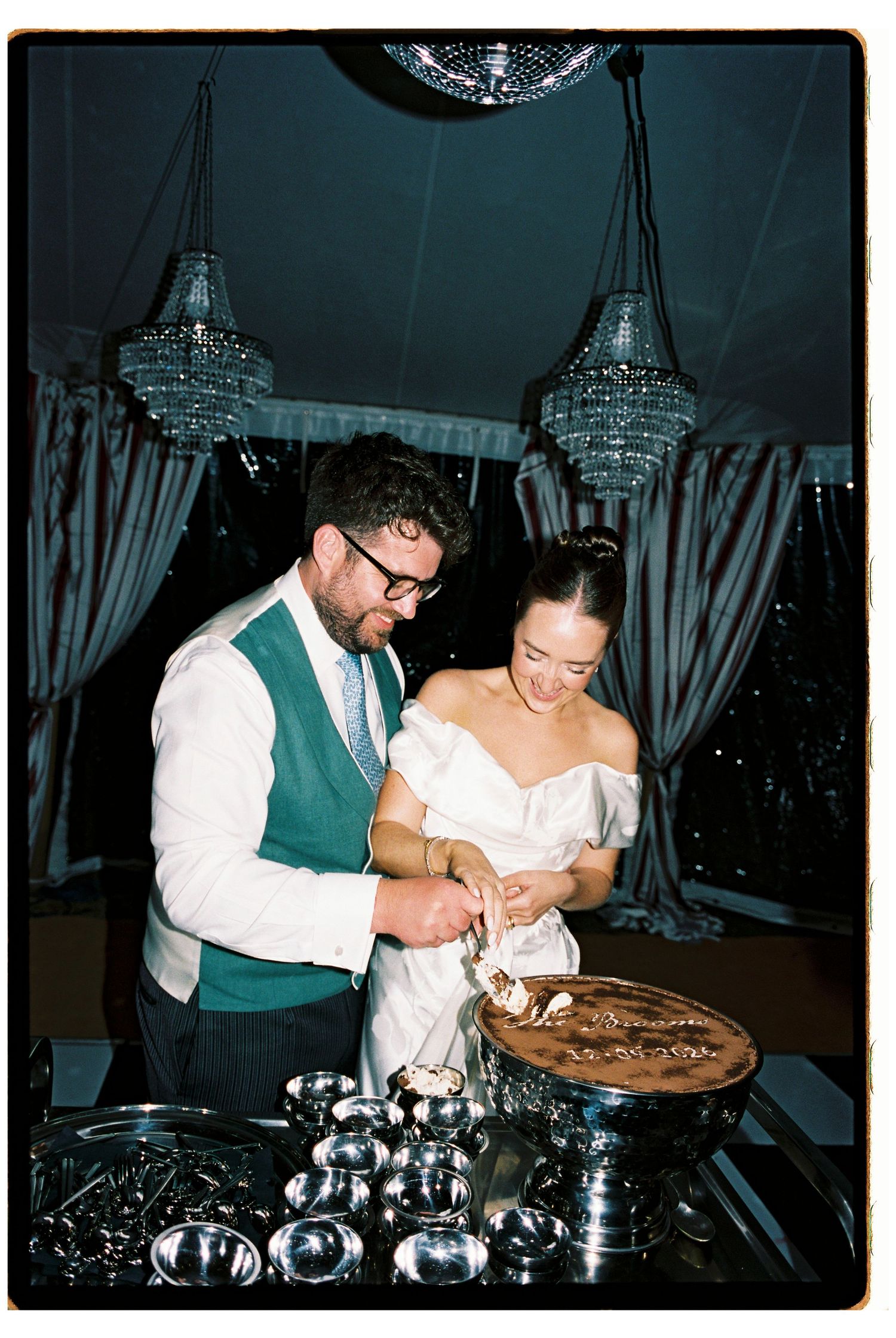 Bride and groom smiling as they cut a chocolate wedding cake together under crystal chandeliers in an elegant tented reception.