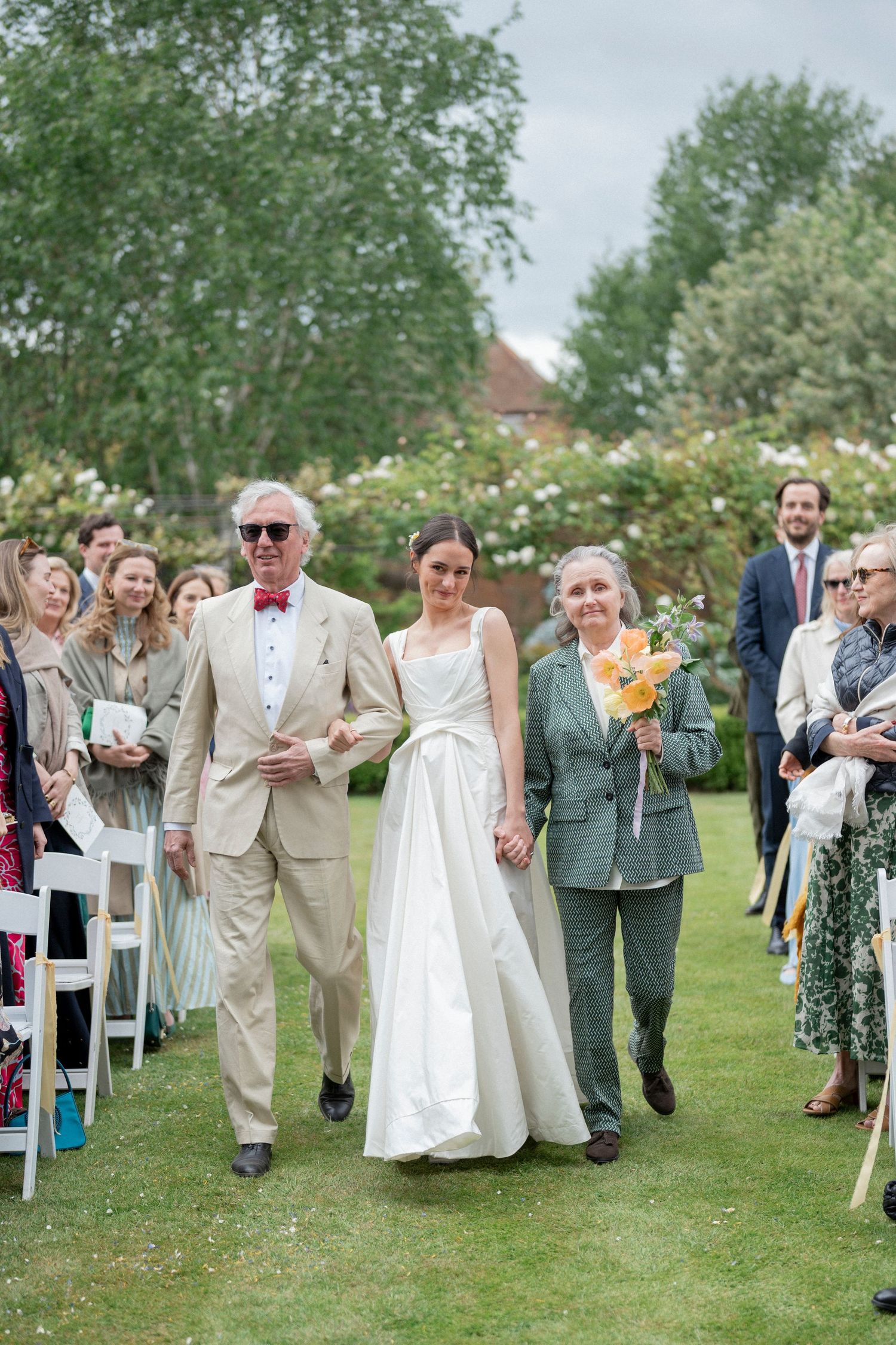 A bride in a white gown walks across a lawn arm in arm with an older man and woman as wedding guests look on.