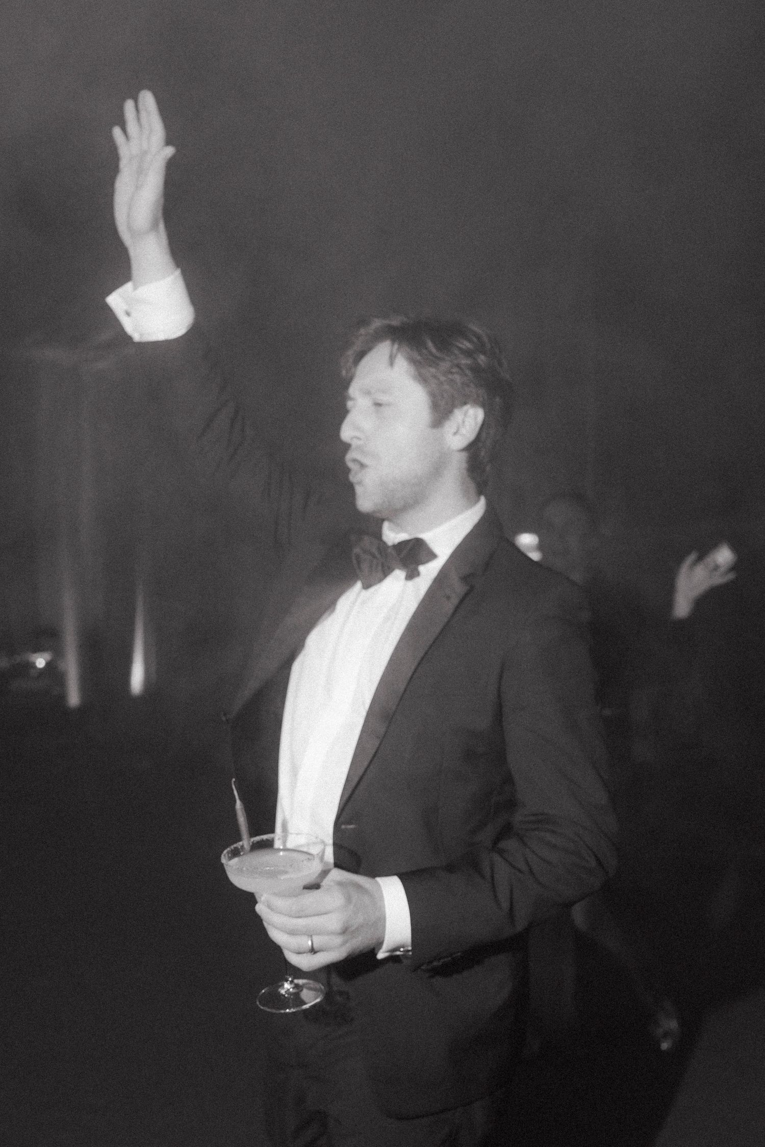 Black and white photo of a man in a tuxedo and bow tie raising one hand while holding a coupe cocktail at a wedding party.
