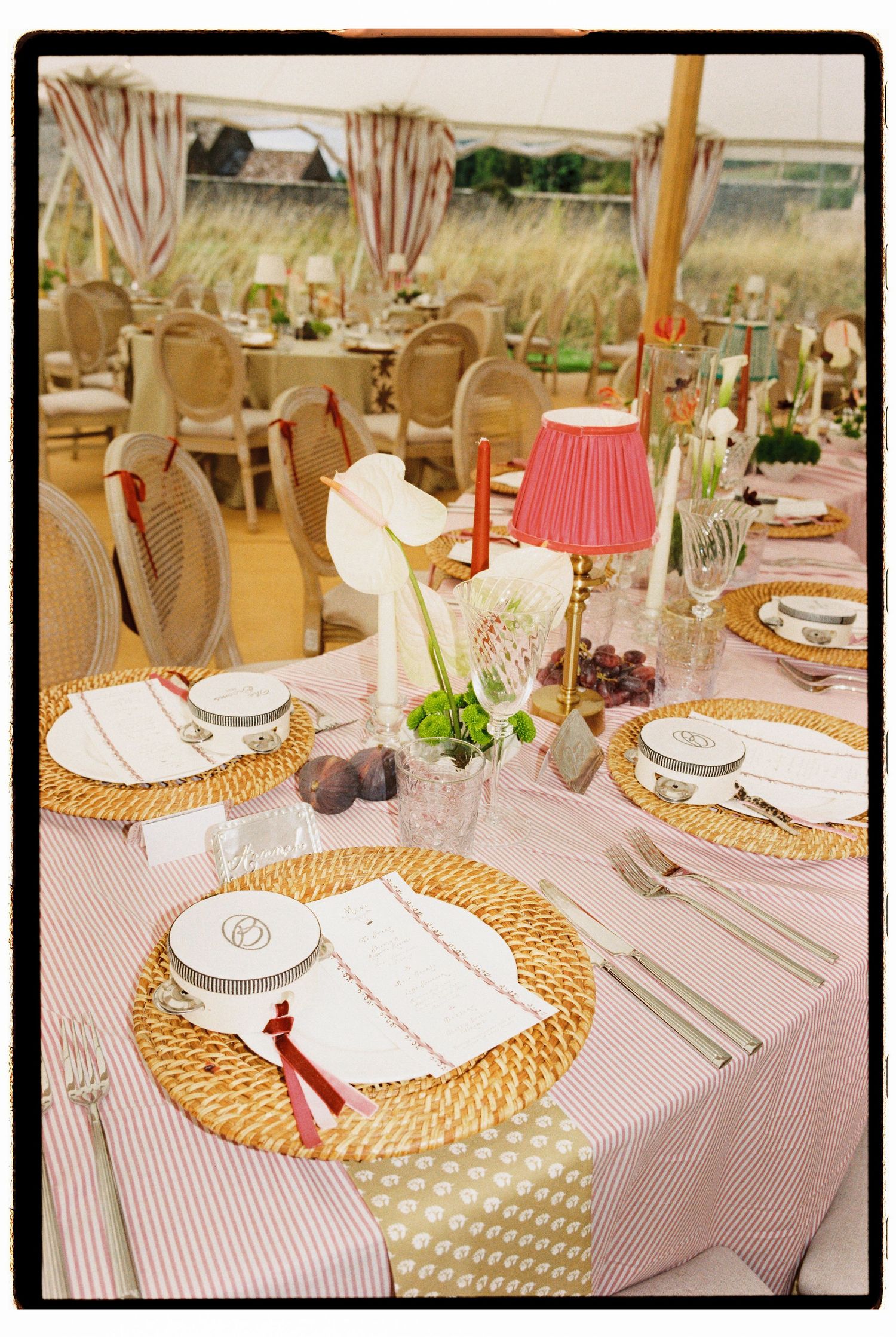 Elegant wedding reception table set with striped linens, wicker chargers, pink lamp, and red candles.