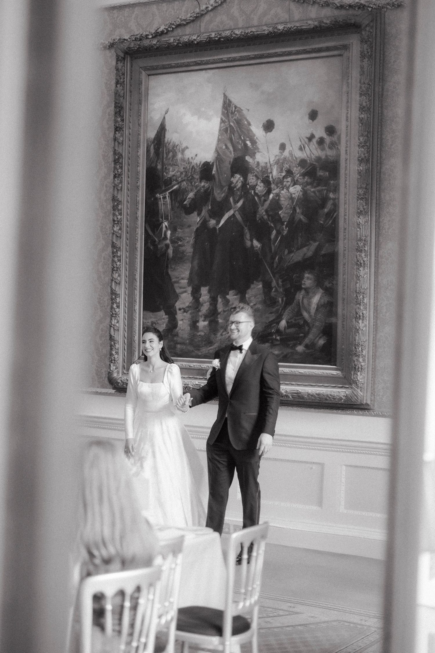 Black and white photo of bride and groom holding hands before a large battle painting in an elegant room.