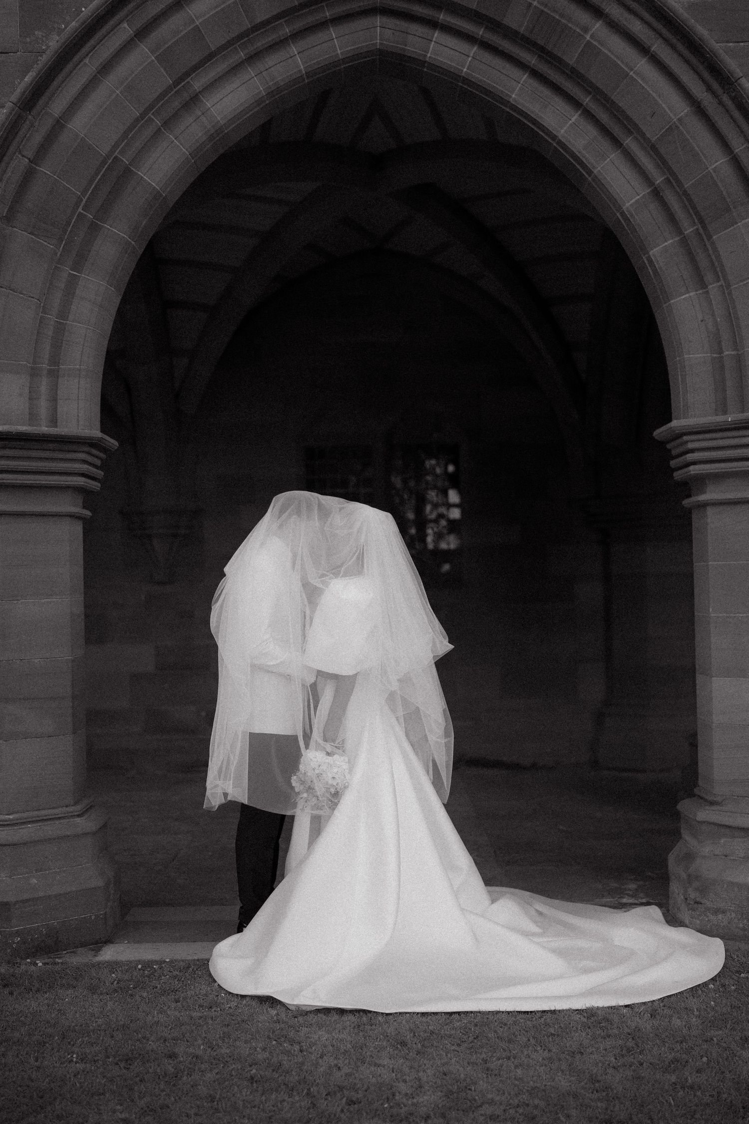 A black and white wedding photo showing a couple in white attire with a flowing veil under a stone archway.
