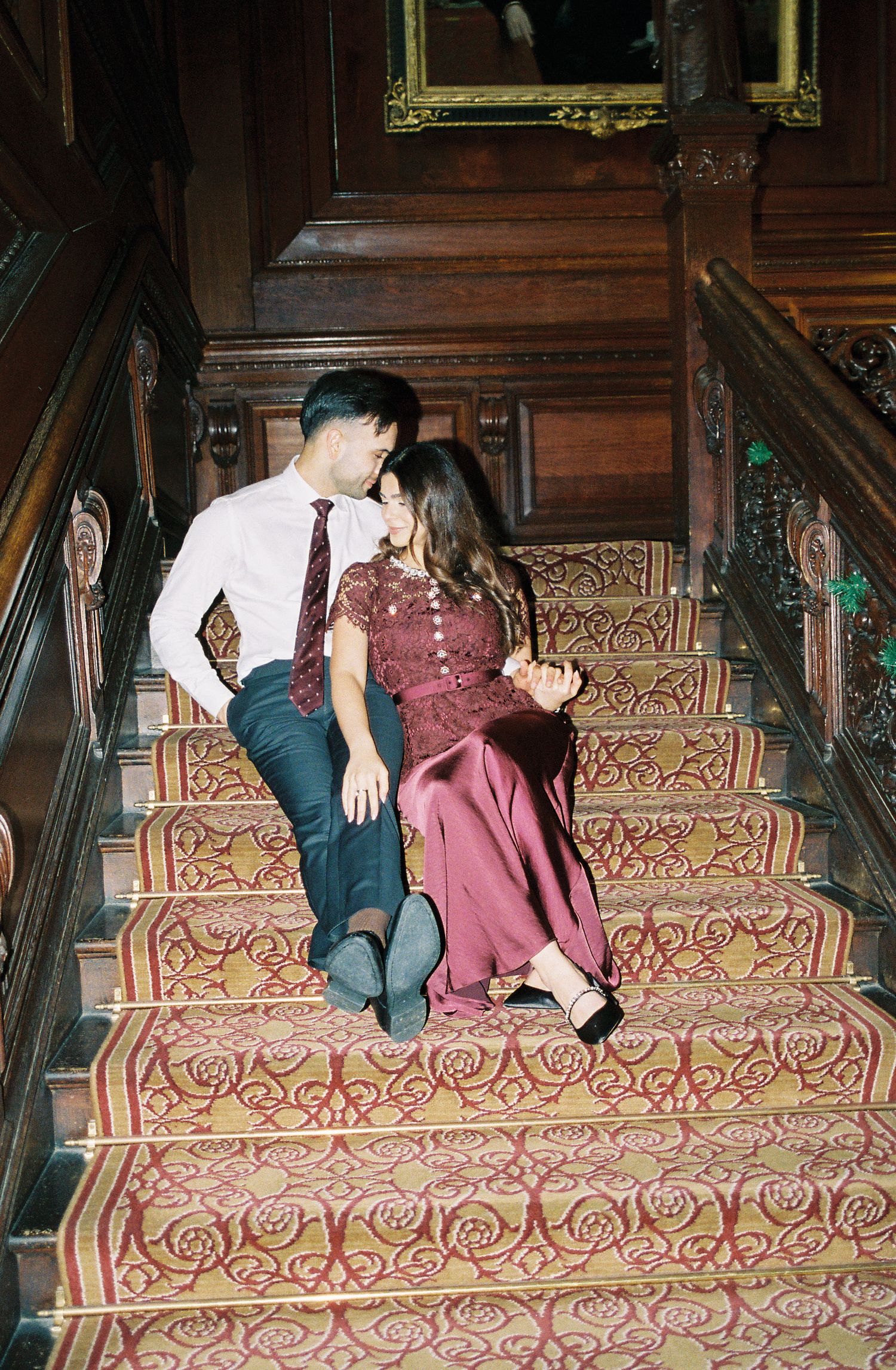 Couple in burgundy outfits sitting romantically on ornate carpeted staircase in elegant wood-paneled mansion.