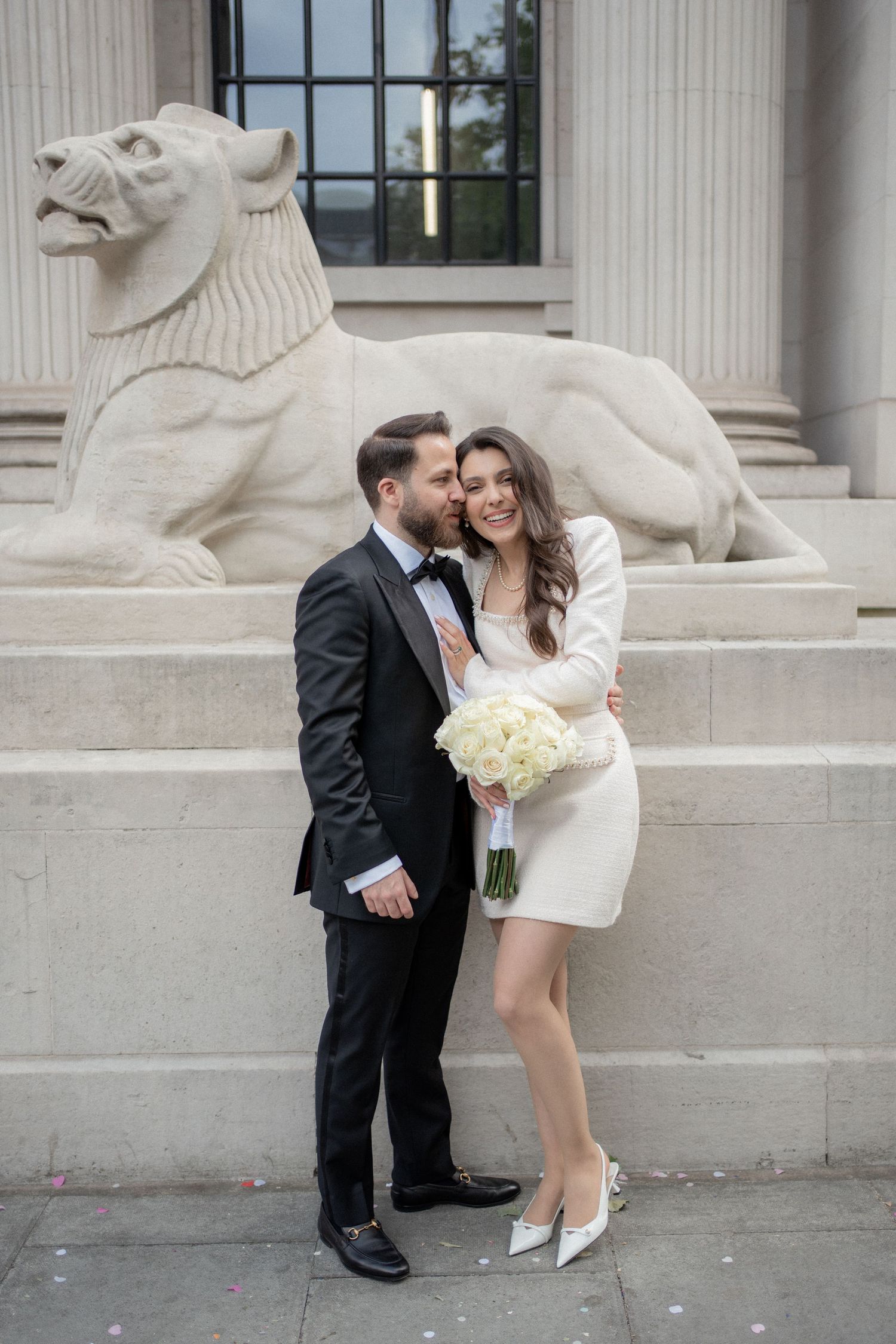 Bride and groom posing in front of a stone lion sculpture, bride holding white bouquet in short wedding dress.