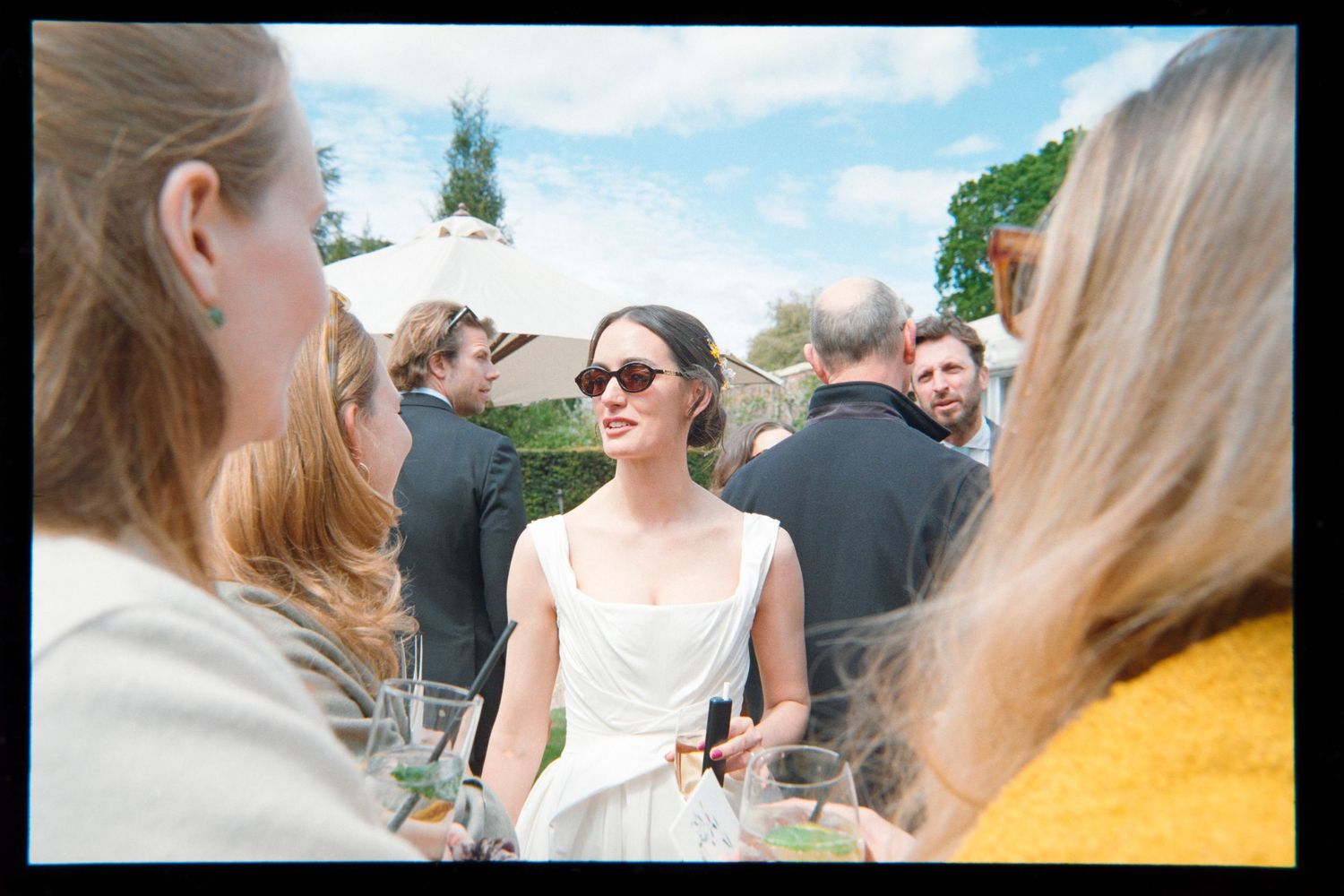 Woman in a white dress and sunglasses stands among wedding guests holding drinks at an outdoor garden party.
