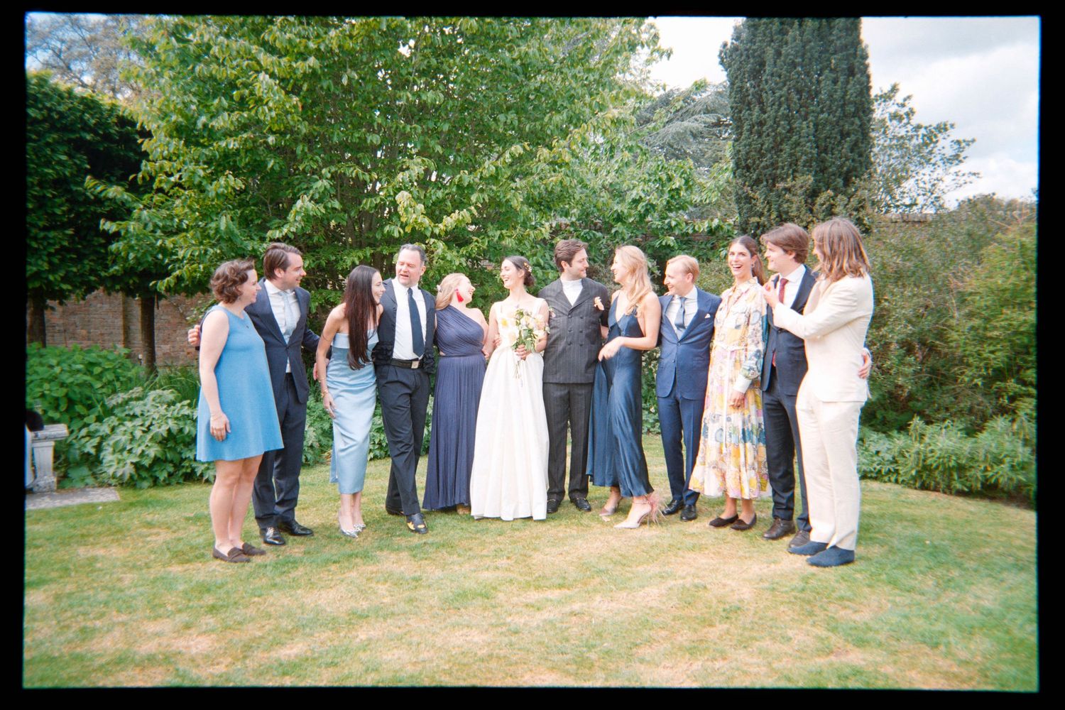 Bride, groom and wedding guests in formal attire pose together for a group photo on a green garden lawn.