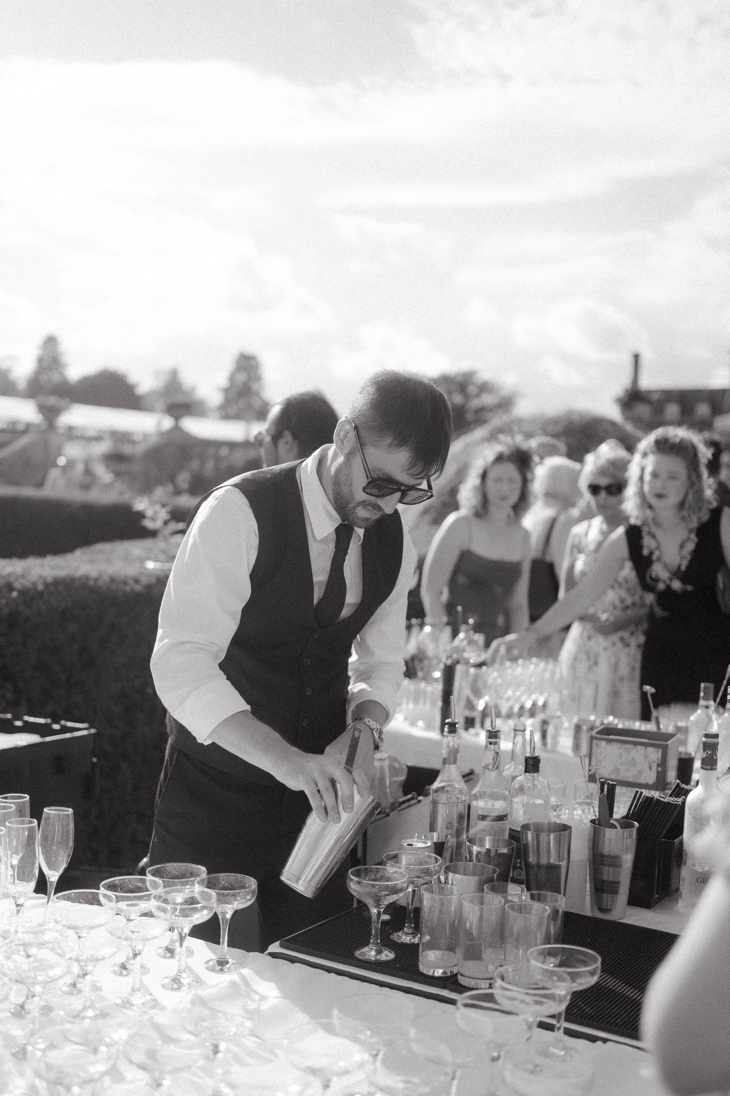 Black and white photo of a bartender pouring cocktails at an outdoor event bar with guests socializing in the background.