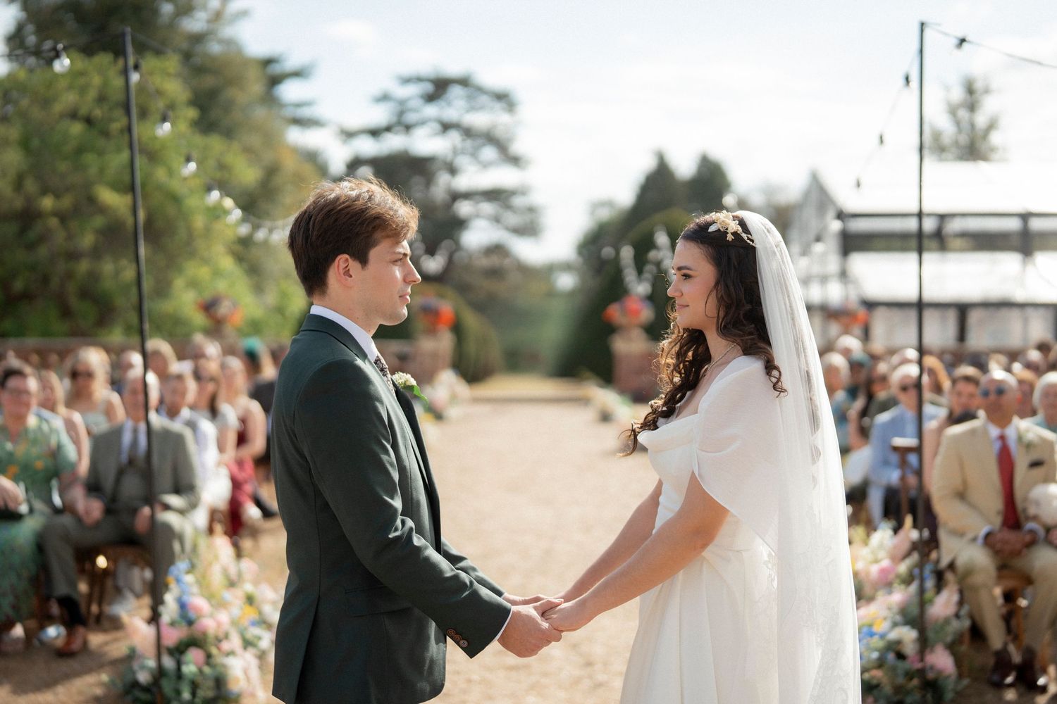 Bride and groom hold hands during outdoor wedding ceremony with seated guests and lush greenery backdrop.