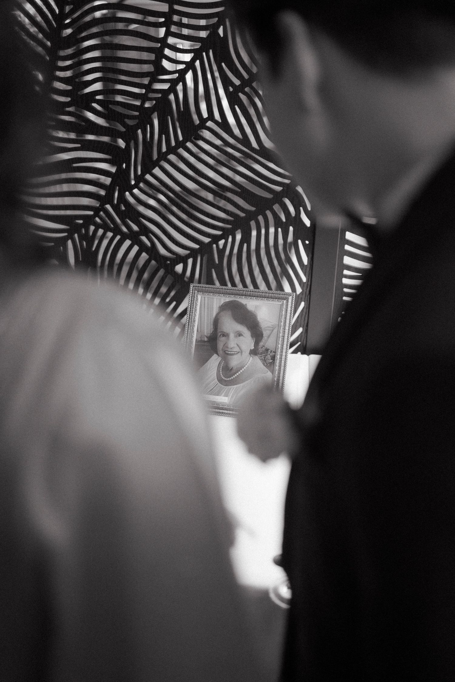 Black and white photo of a framed portrait of a woman with pearls, viewed between blurred figures at a memorial.