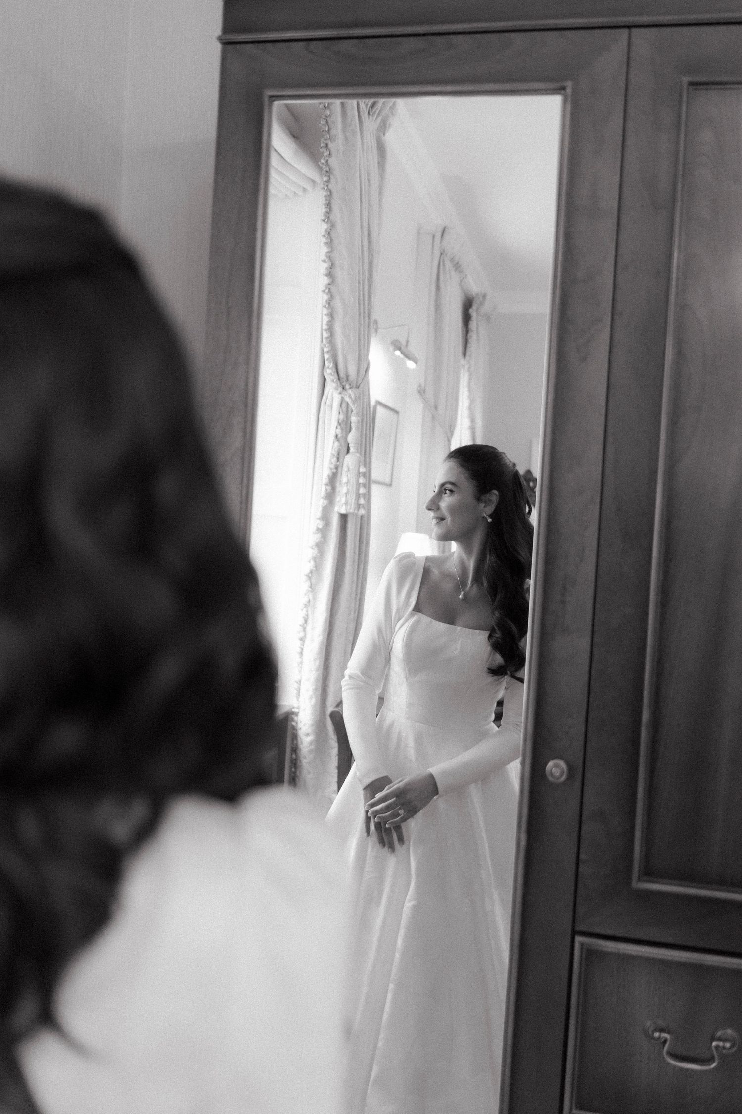 Black and white photo of a bride in a white gown admiring her reflection in a mirror on her wedding day.