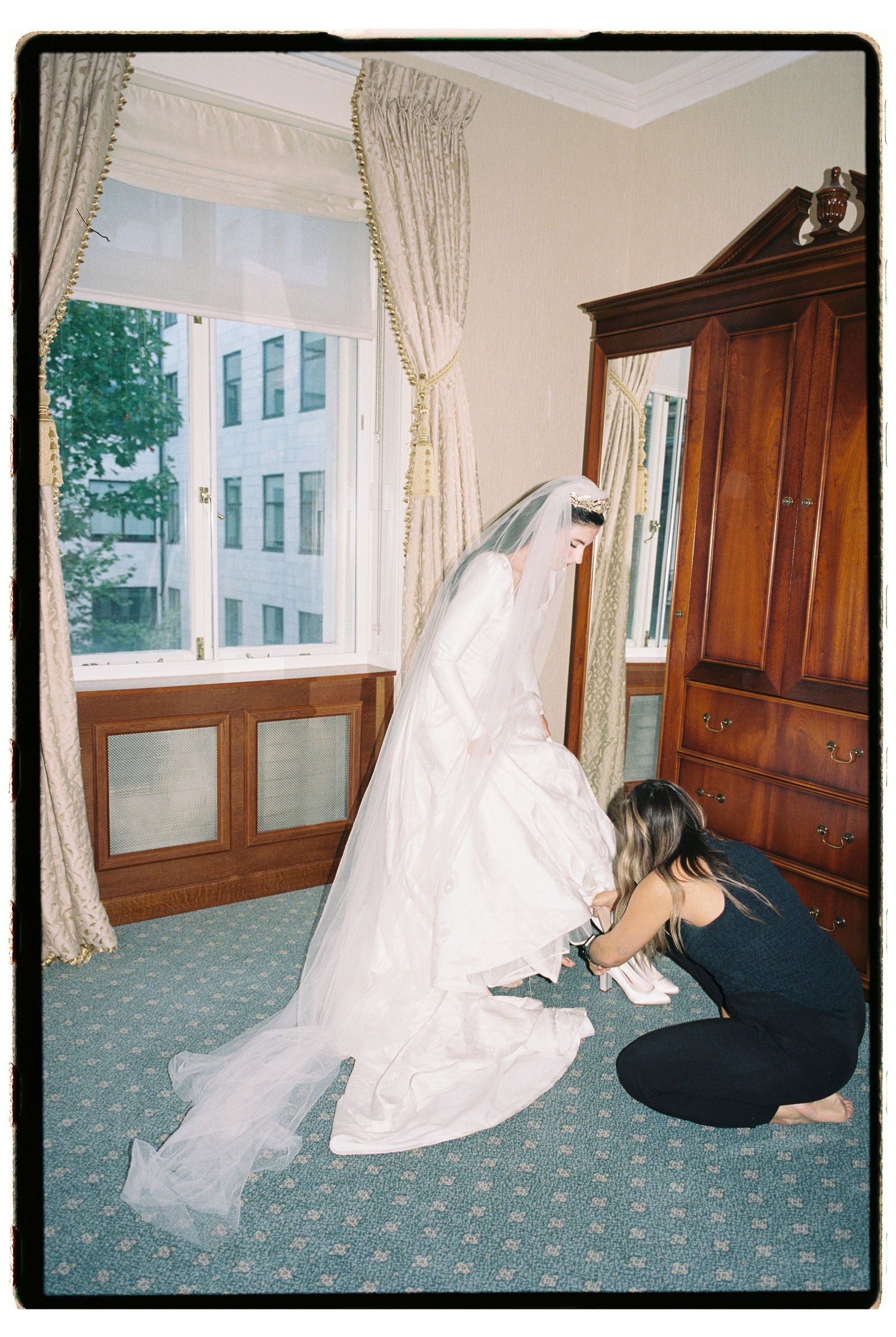 A bride in a white gown and veil stands while an assistant adjusts her dress in an elegant hotel room.
