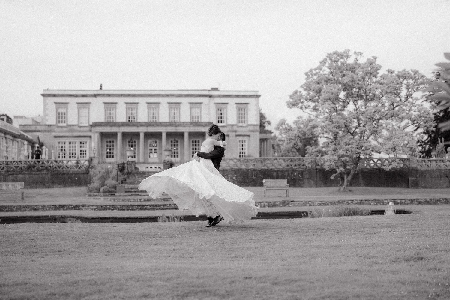 A black and white photo of a bride twirling in her dress on a grand estate lawn.