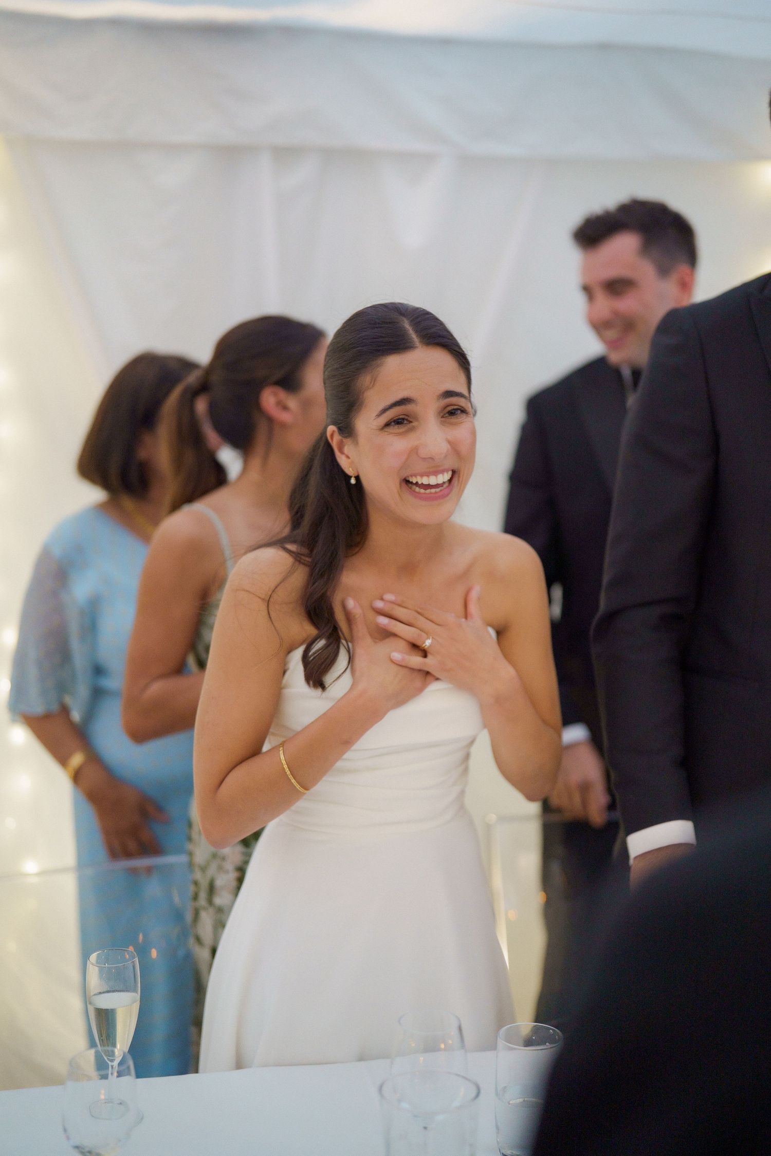 A bride in a white halter dress laughs joyfully during wedding reception speeches.