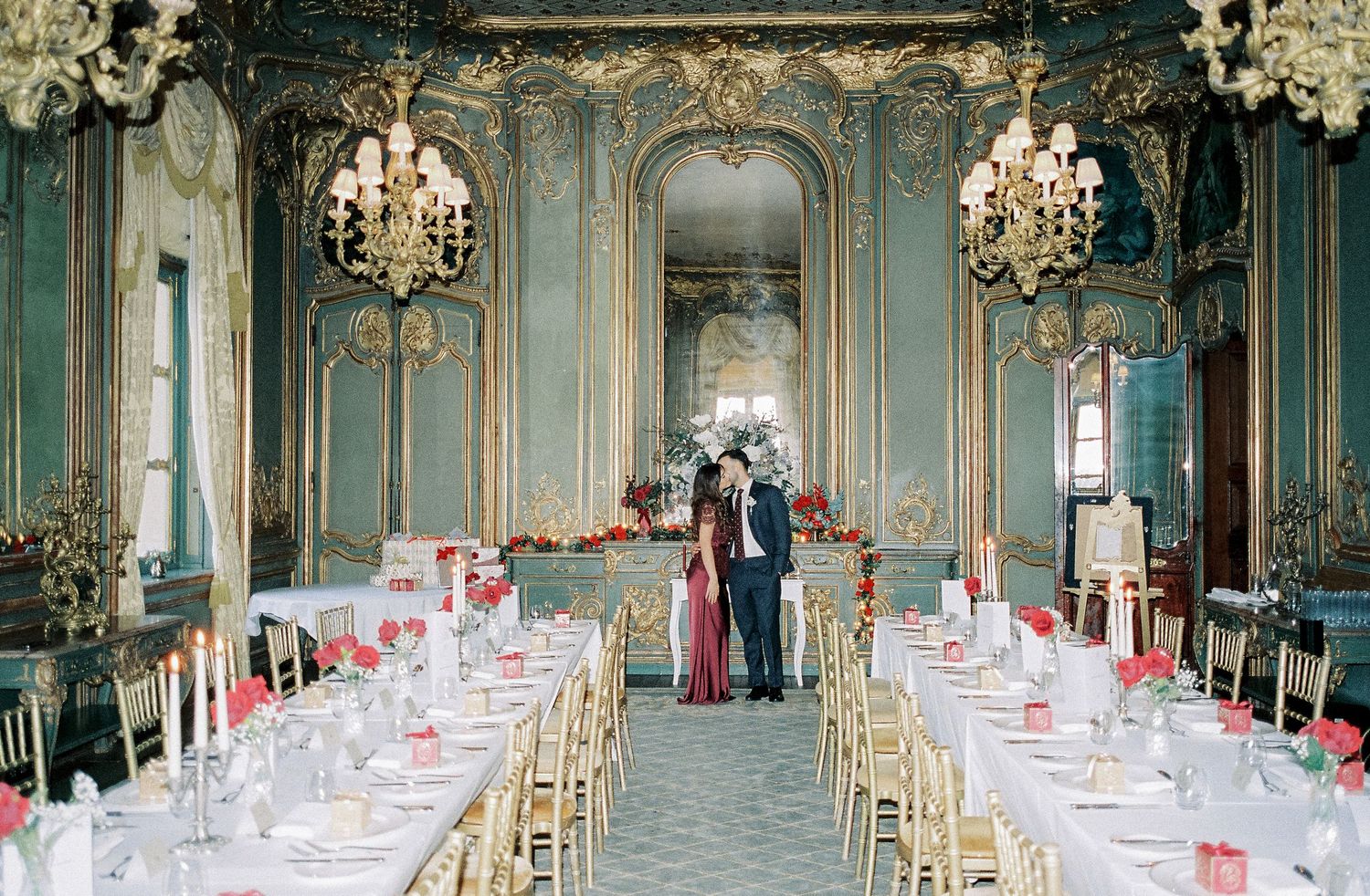 Elegant wedding reception in ornate blue and gold ballroom with chandeliers, long tables, and couple standing at altar.