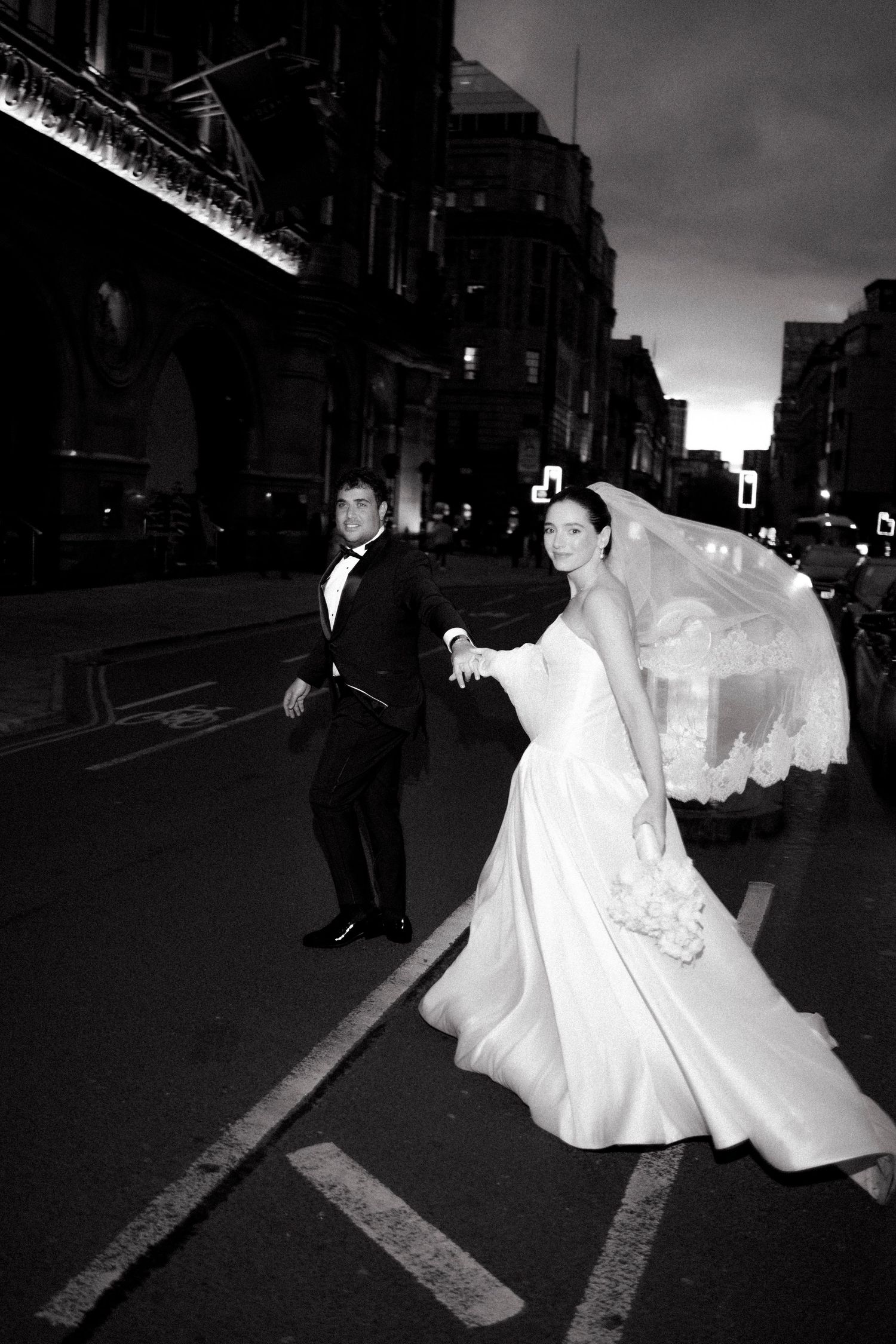 Black and white photo of a bride and groom holding hands and crossing a city street at dusk, her long lace veil flowing behind her.