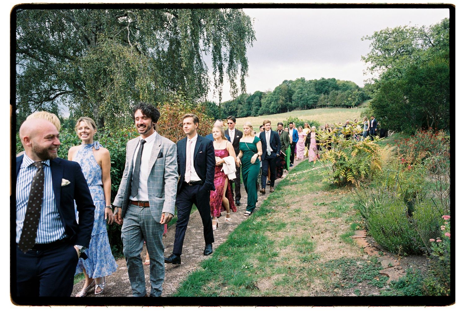 Wedding guests walking in a line along a garden path surrounded by lush greenery at an outdoor celebration.