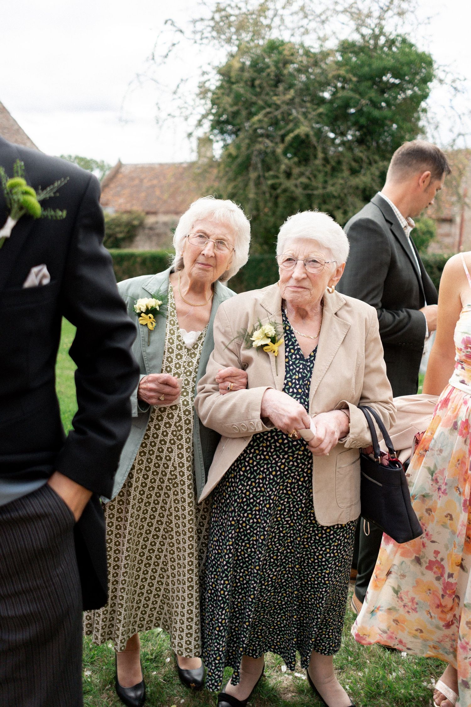 Two elderly female wedding guests with corsages smiling and holding hands at outdoor garden wedding.