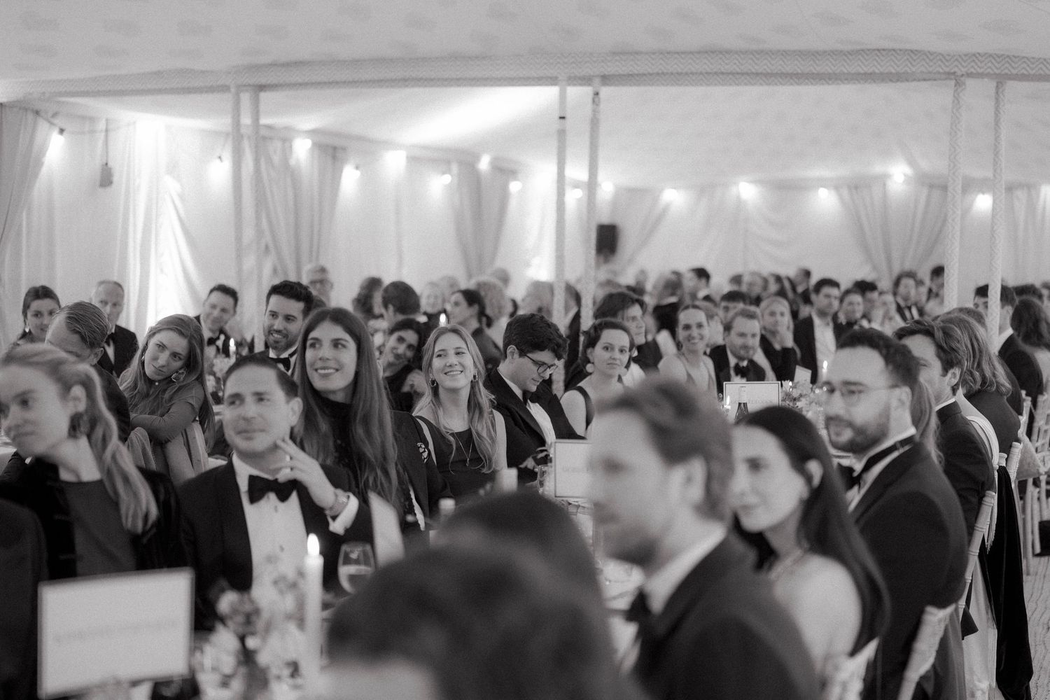 Wedding guests in formal attire smile and chat at long tables inside a softly lit reception marquee in black and white.