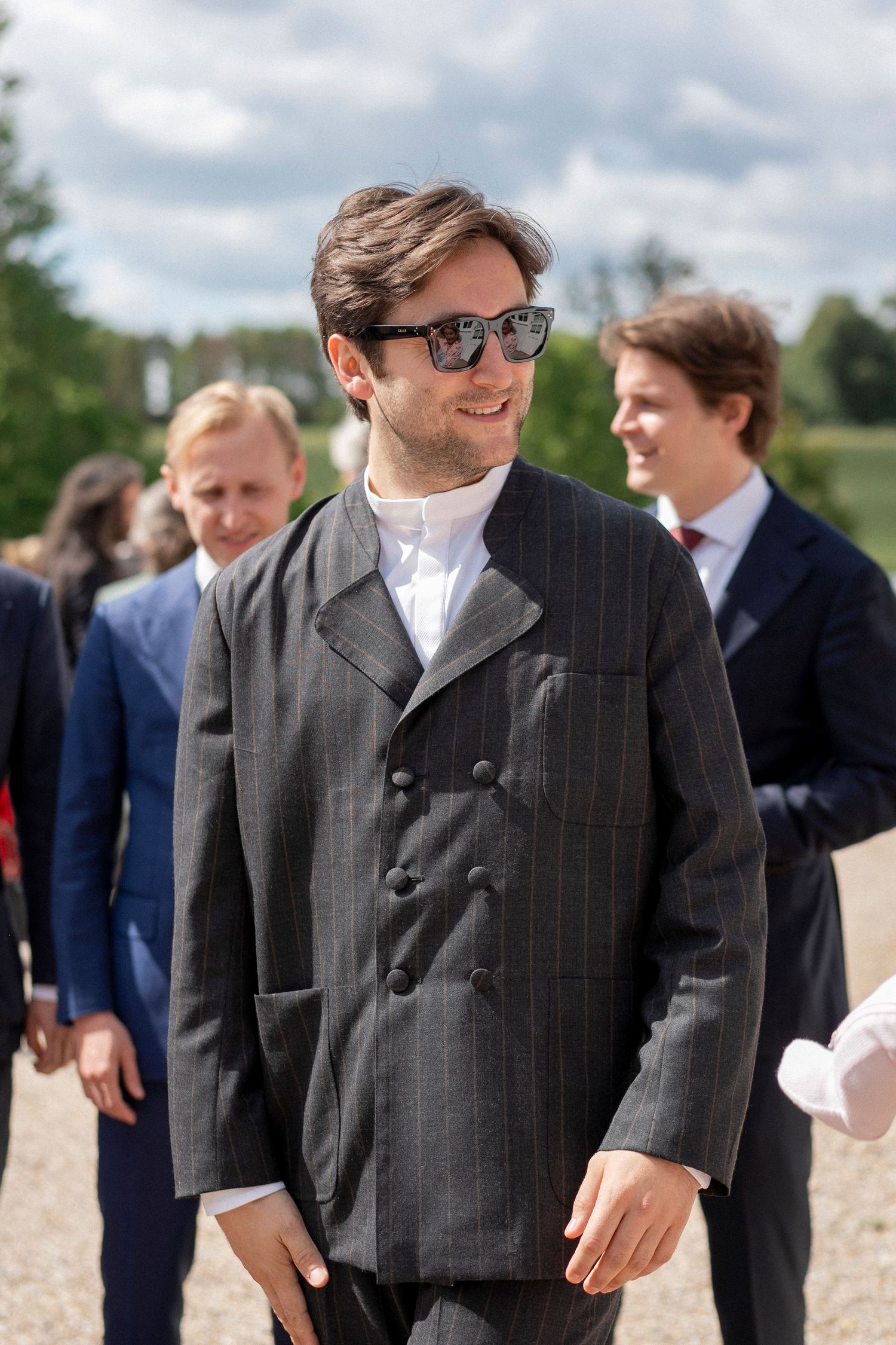 Smiling man in sunglasses and a pinstripe double-breasted jacket stands among wedding guests outdoors on a cloudy day.