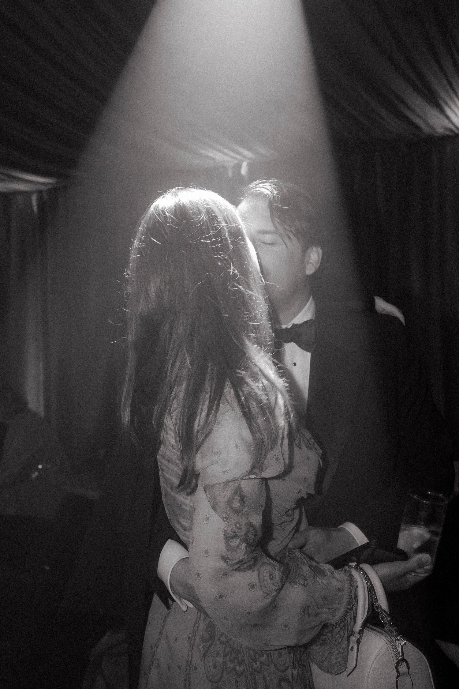 Black and white photo of a couple embracing in a beam of light, she wears a floral dress, he wears a tuxedo and bow tie.